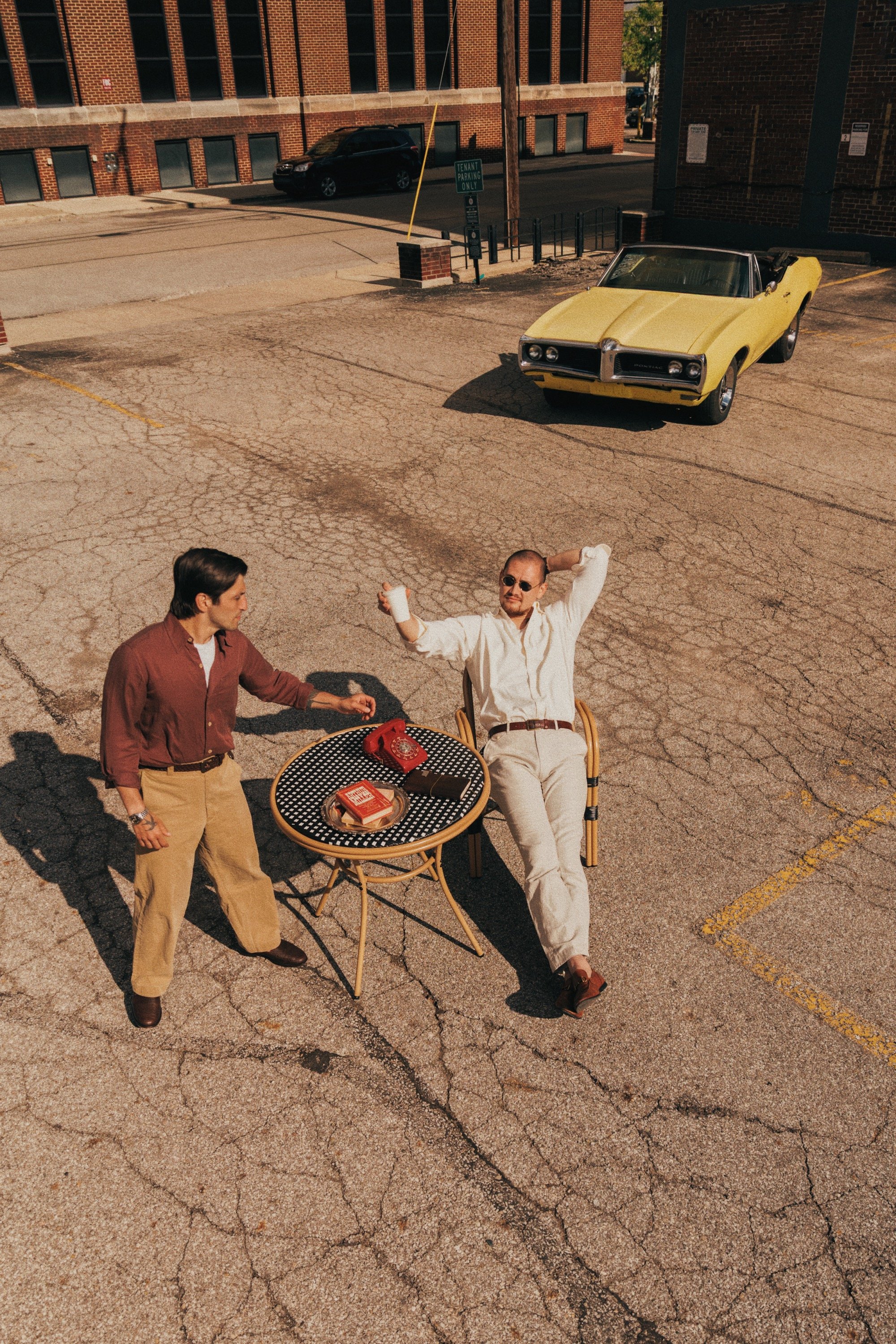 Two men are sitting and standing in an empty parking lot, one is holding a white cup, and the other has his hands behind his head. A yellow classic car is parked nearby, with a table with a telephone and some books on it in front of them. Background 
