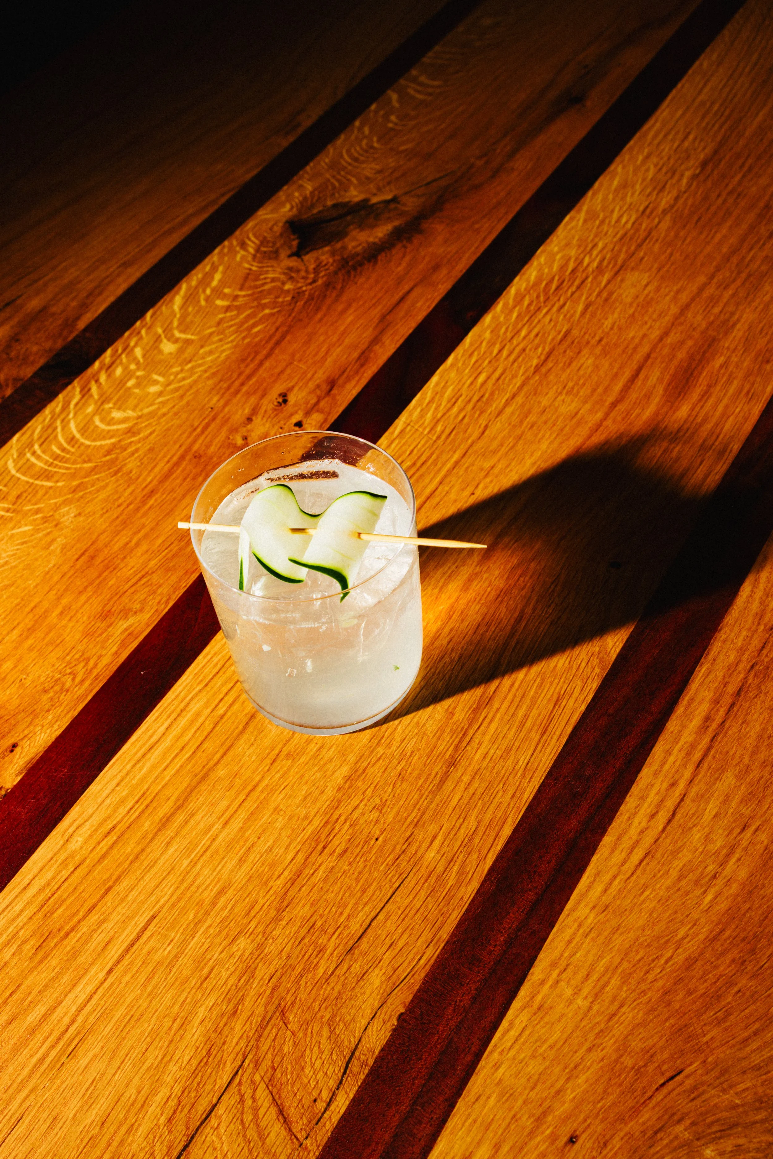 A clear glass with a cloudy drink garnished with a cucumber peel, placed on a wooden table with warm tones and dark inlays.