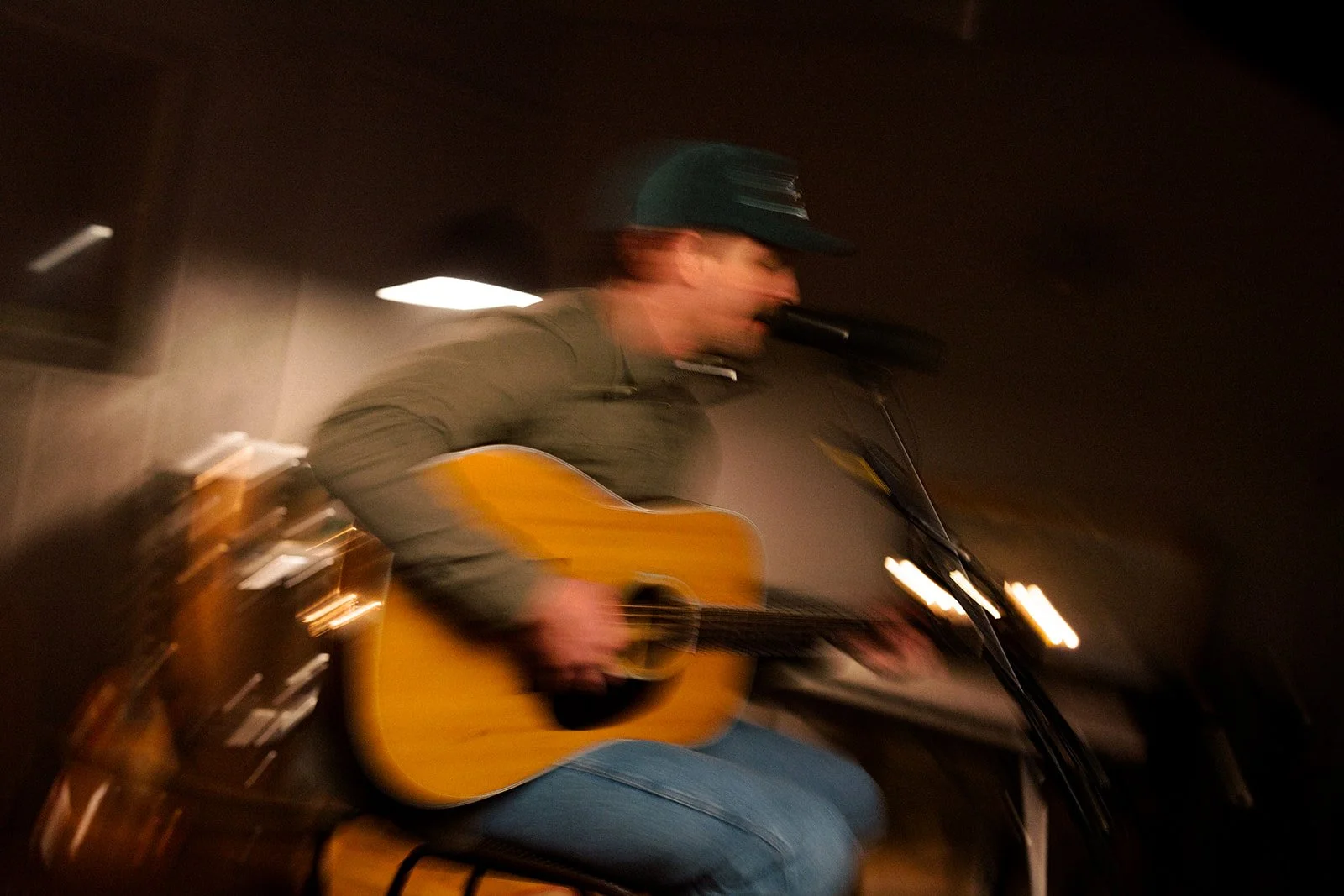 A person wearing a hat playing an acoustic guitar and singing into a microphone in a dimly lit room.