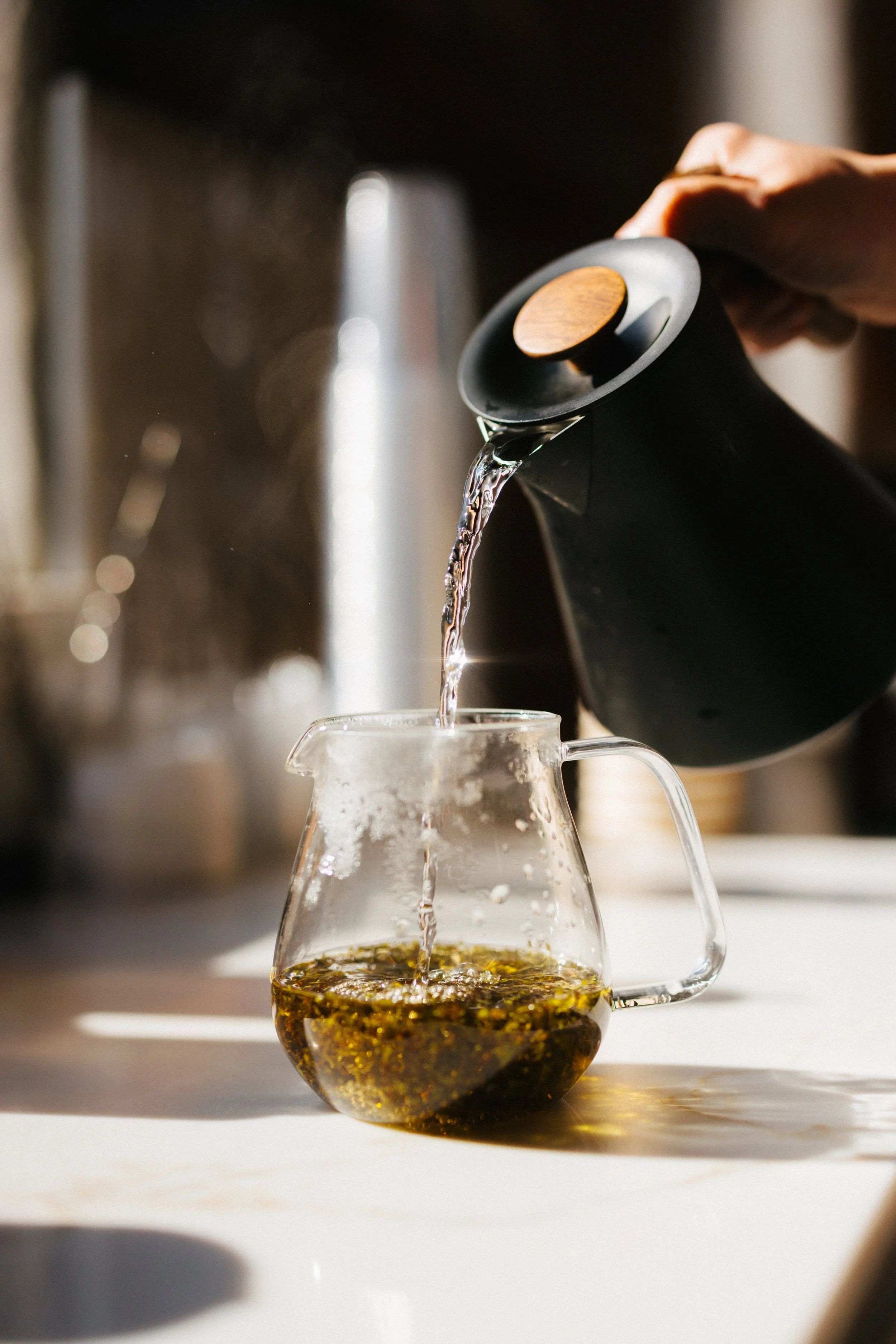 Close-up of a person pouring hot water from a black kettle with a wooden handle into a glass pitcher filled with coffee grounds.