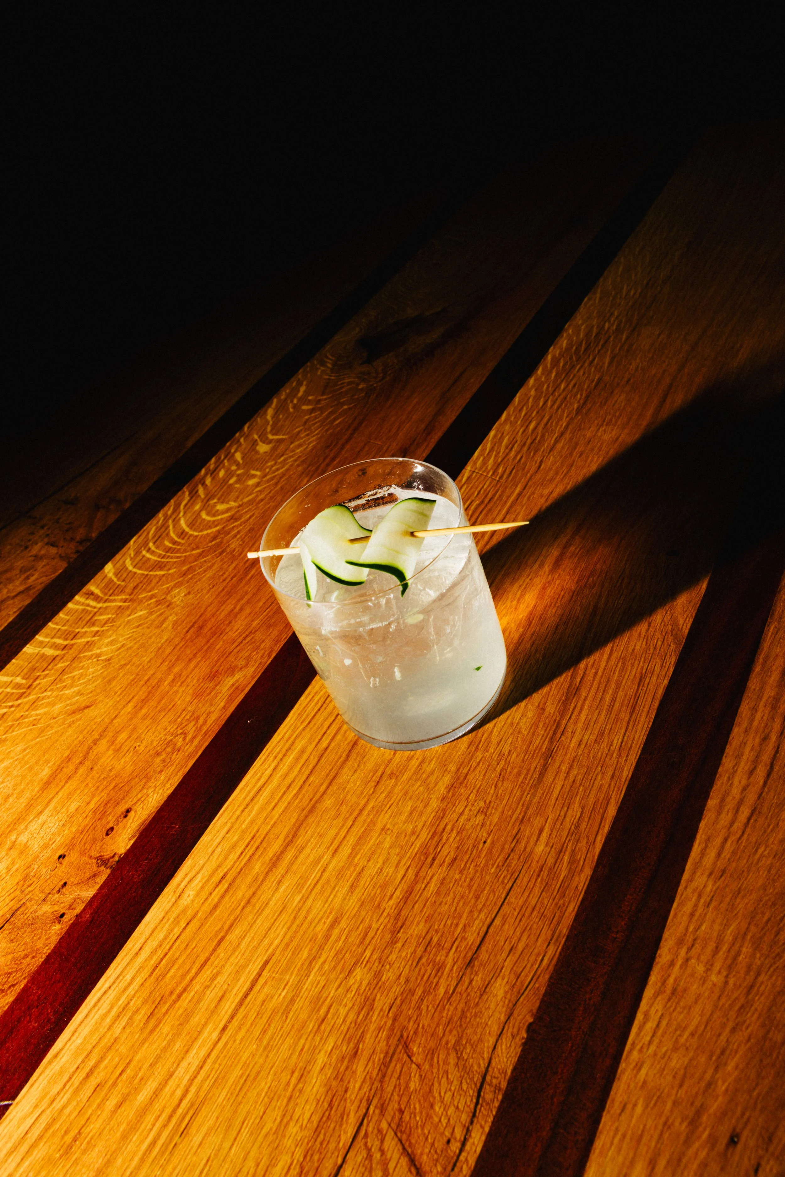 A glass with a clear beverage garnished with cucumber slices on a wooden surface with warm lighting.