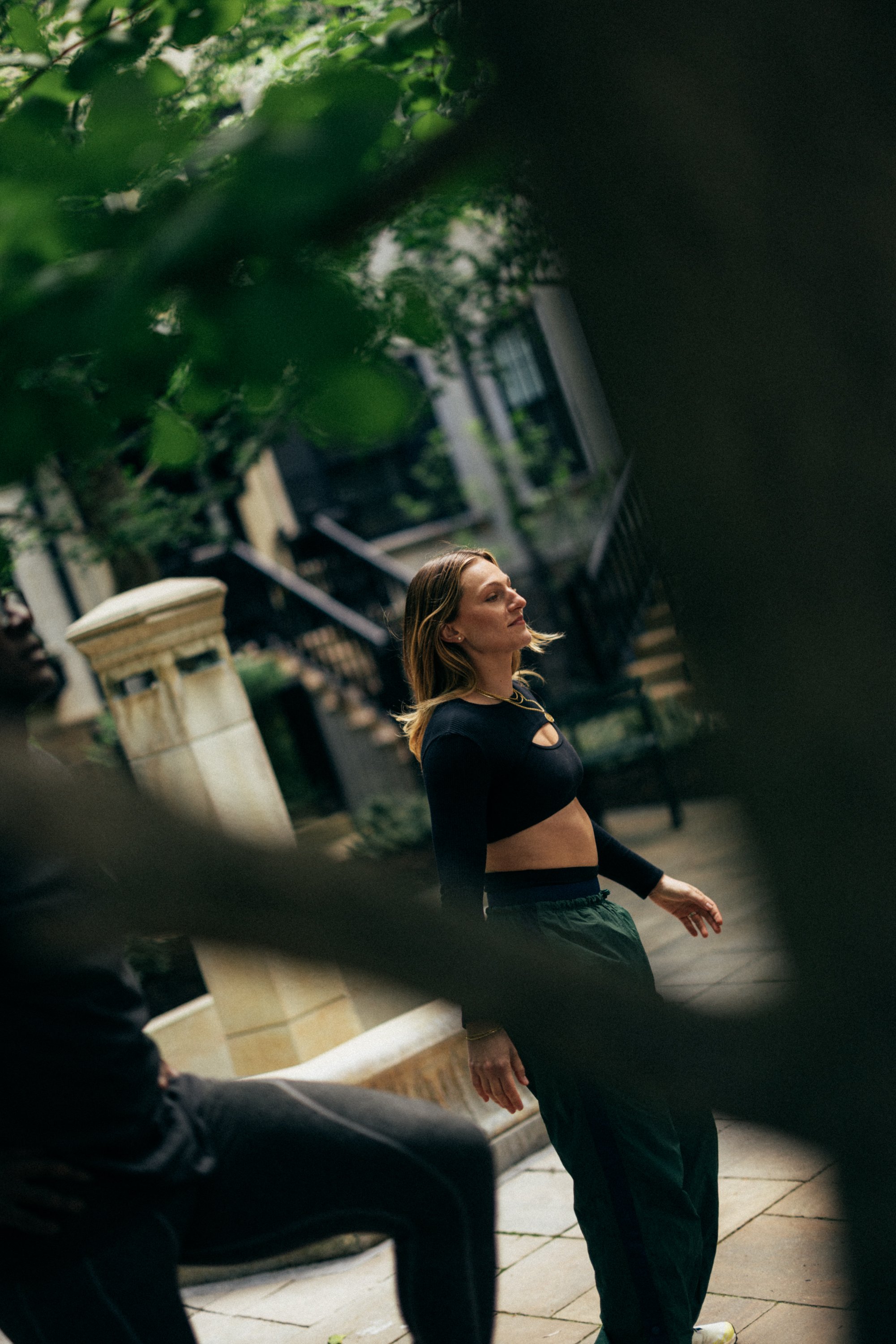 A woman with blonde hair styled in loose waves, wearing a black long-sleeve crop top and dark sweatpants, stands outdoors near a staircase and railing in a city setting during the daytime. Green foliage partially obscures the view in the foreground.