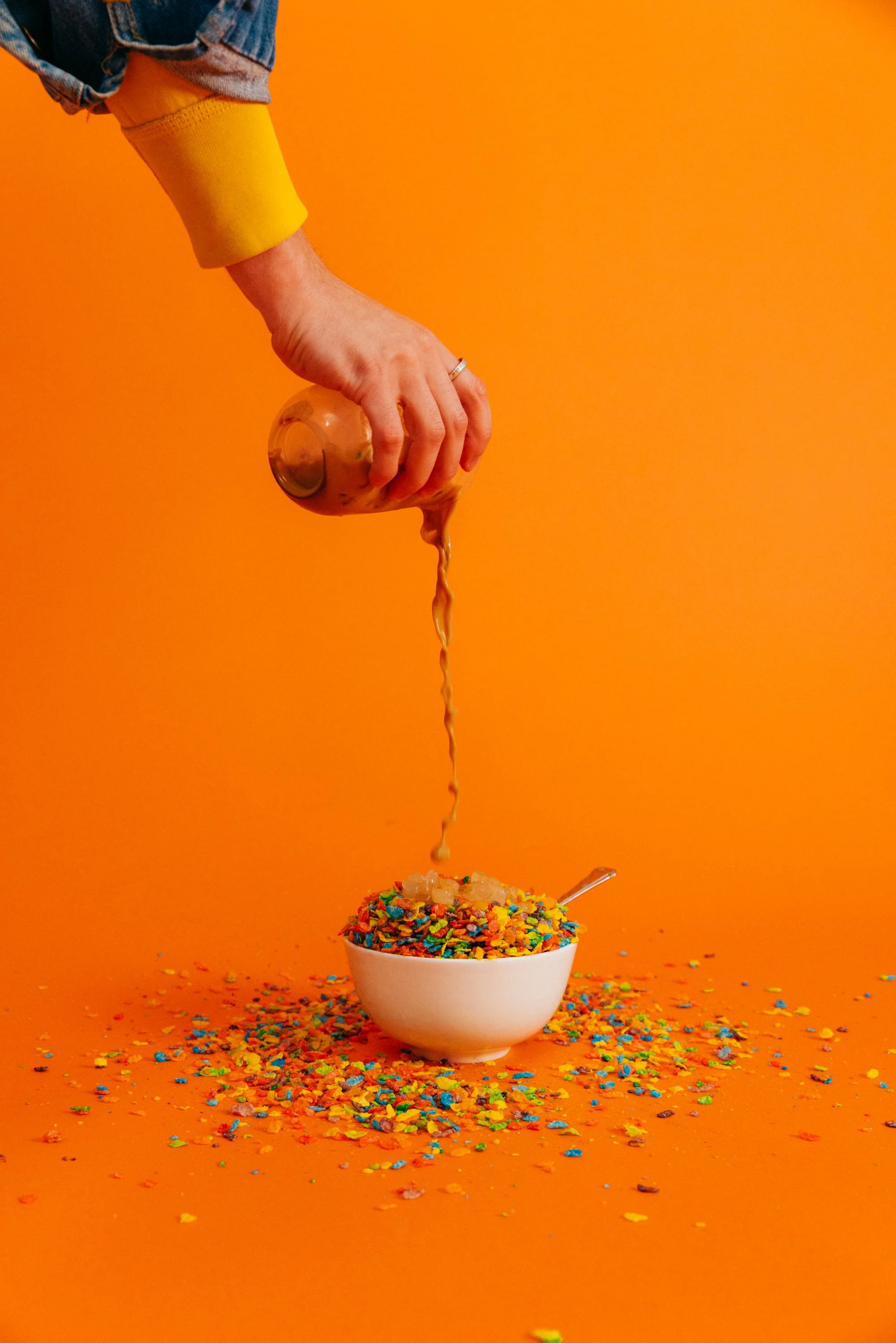 A hand pouring colorful sprinkles from a cup onto a bowl of cereal against an orange background.