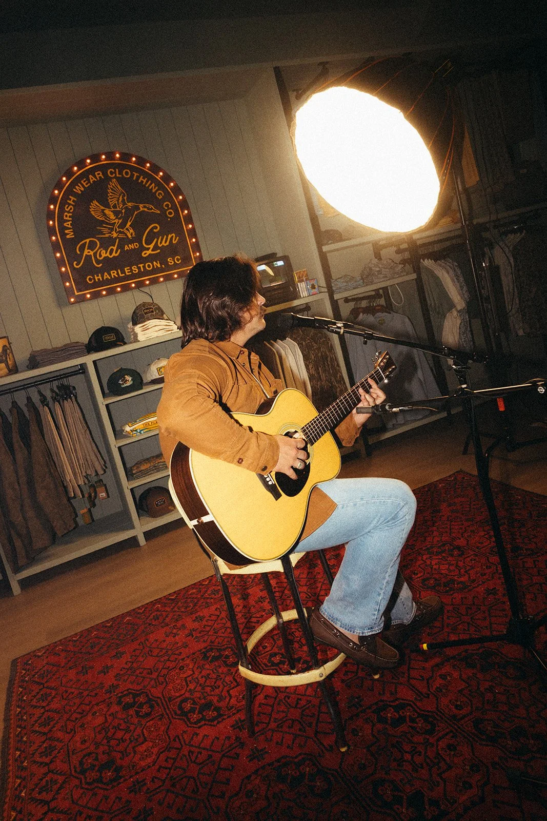 A man with dark hair playing an acoustic guitar while singing into a microphone. He is sitting on a round stool on a red patterned rug inside a clothing store with a sign that reads "Marsh Wear Clothing Co." and hats on display in the background.