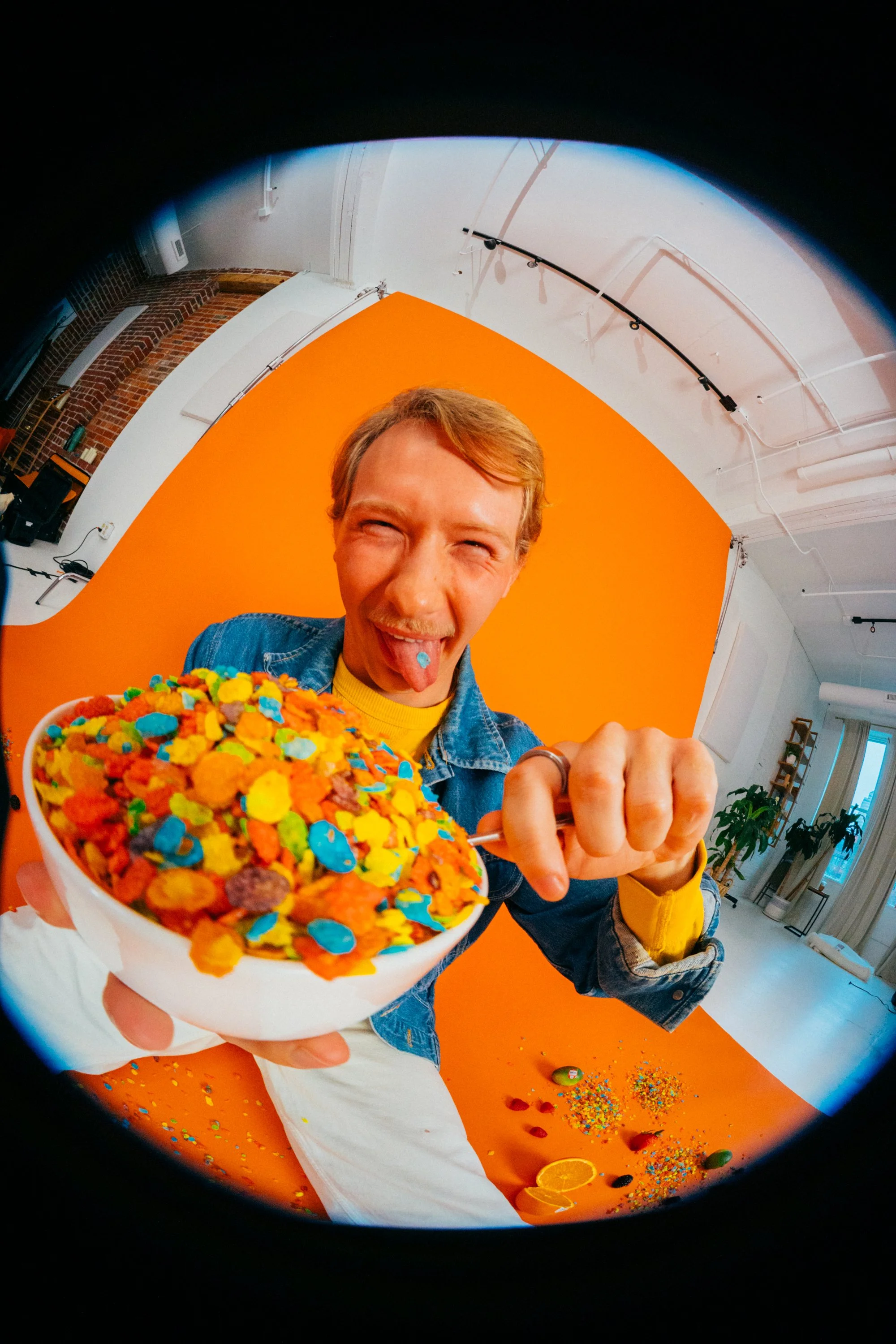 Person with short blonde hair and a denim jacket sticking out their tongue, holding a bowl of colorful cereal with rainbow sprinkles against an orange background in a studio setting.
