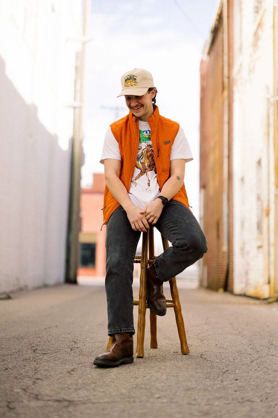 A young man sitting on a wooden stool in an alleyway, smiling and looking down, wearing a beige cap, orange vest, graphic T-shirt, black jeans, and brown boots.
