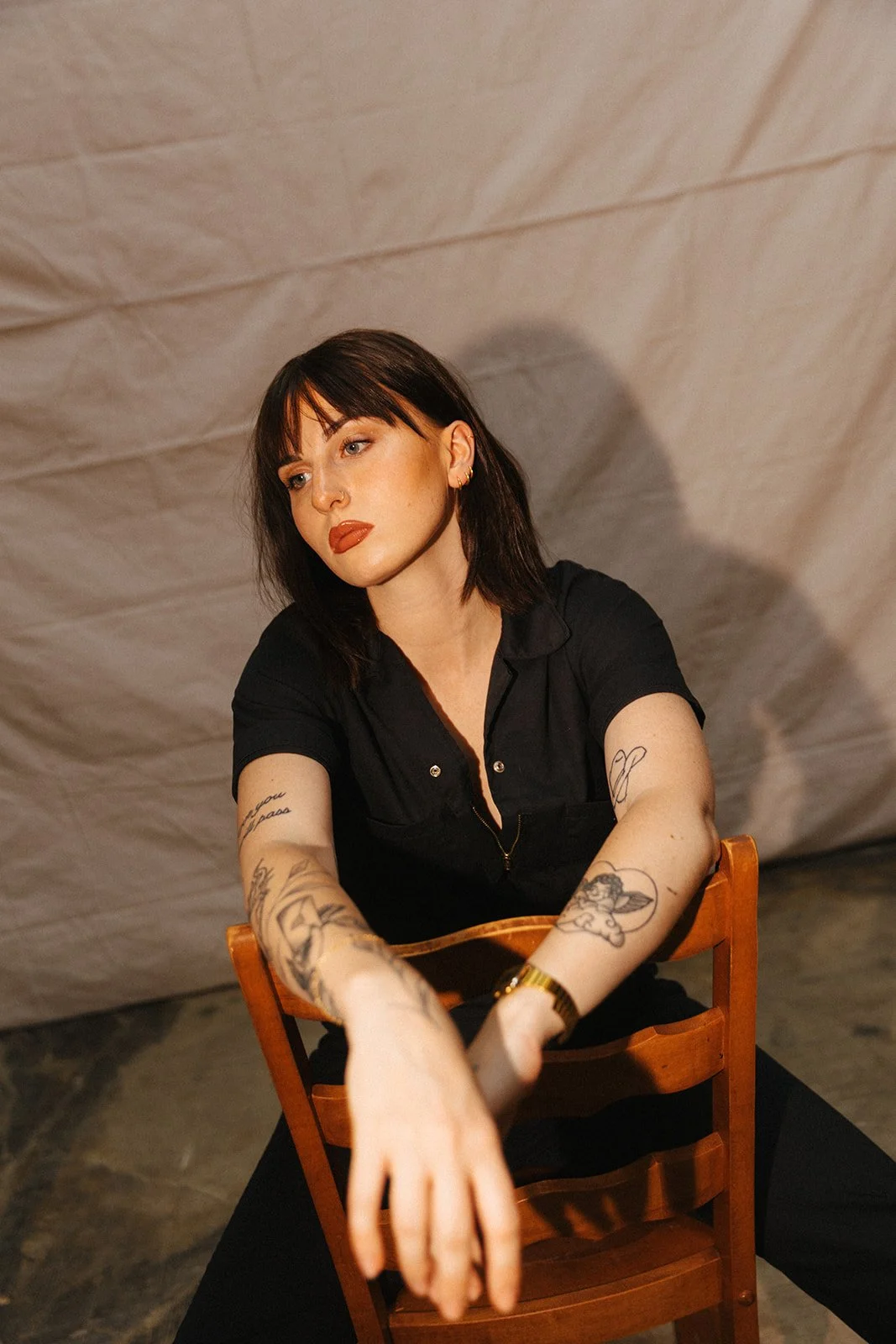 A woman with dark hair and tattoos sitting on a wooden chair against a beige background, wearing a black top and gold watch.