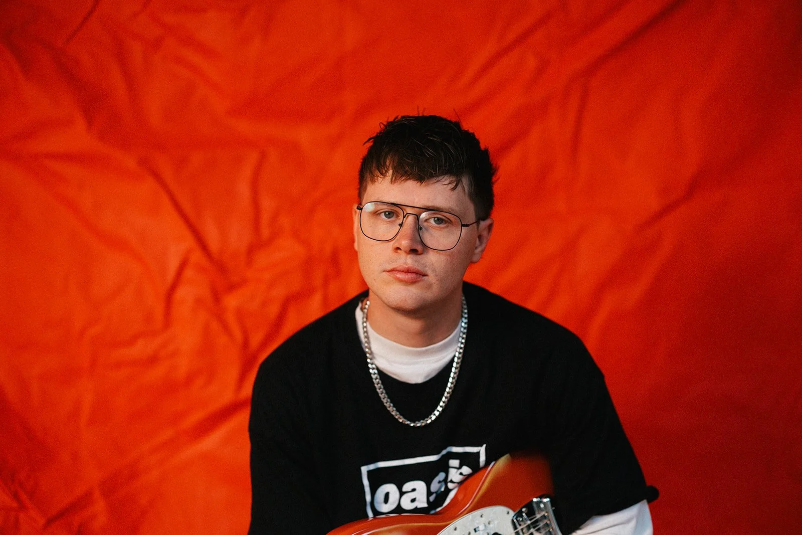 A young man with short dark hair and glasses wearing a black shirt, a silver chain, and a white long sleeve shirt underneath, holding an electric guitar, against a textured orange background.
