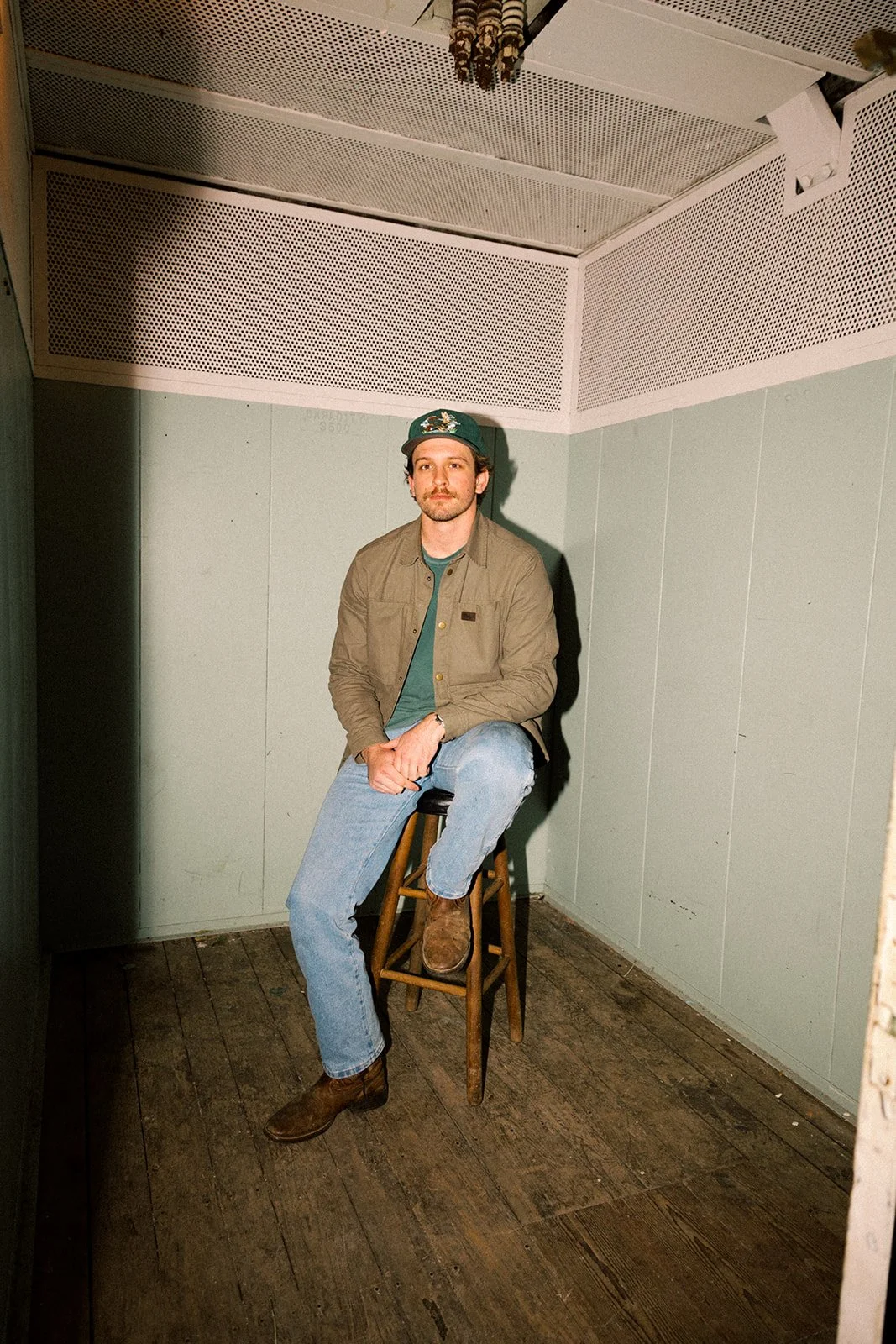 A man sitting on a wooden stool in a small, plain room with light green walls and a wooden floor, wearing a baseball cap, khaki jacket, blue jeans, and brown boots.