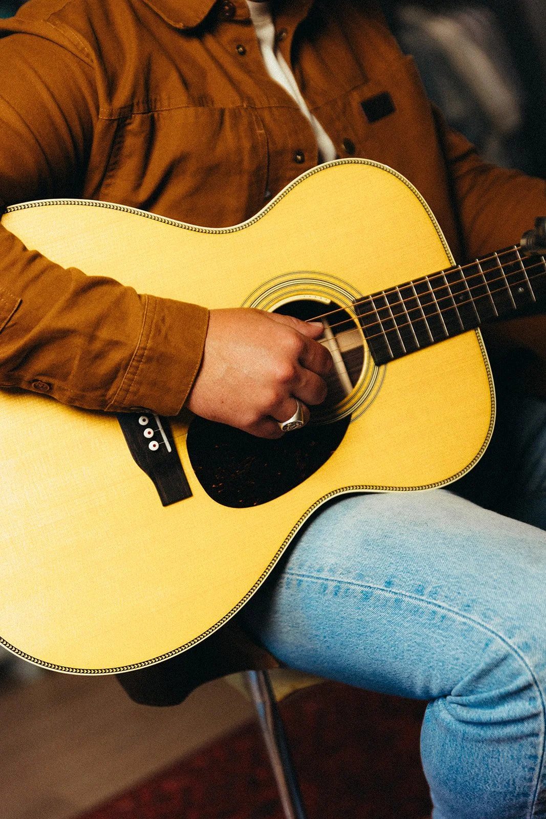 Person playing an acoustic guitar, wearing a brown jacket and light blue jeans.