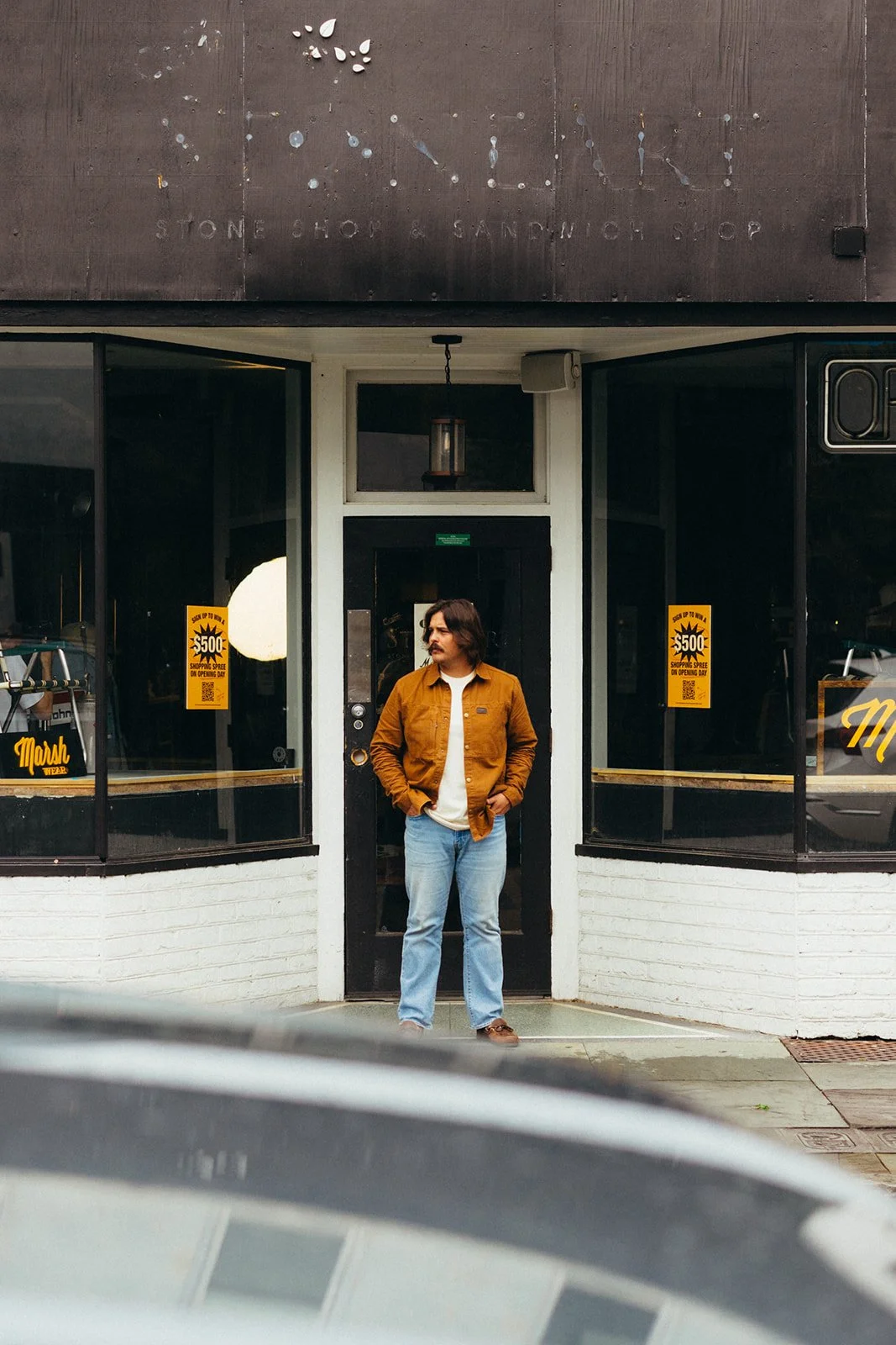 A man with long hair and a mustache wearing a brown jacket, white shirt, and jeans, standing in front of a store entrance with black and white walls, window signs, and a hanging light fixture.