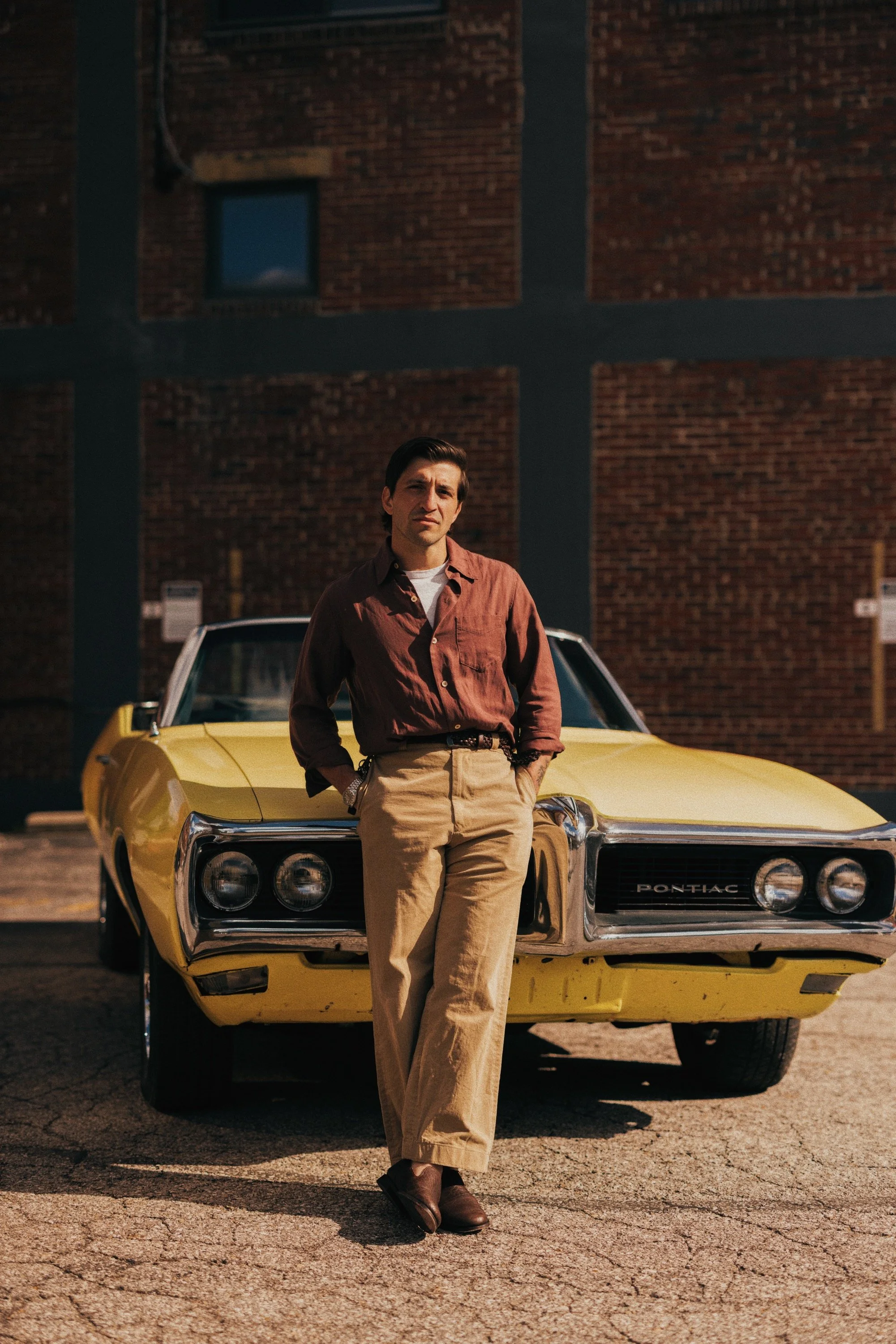 A man in a brown shirt and beige pants standing in front of a yellow Pontiac car parked outside a brick building.