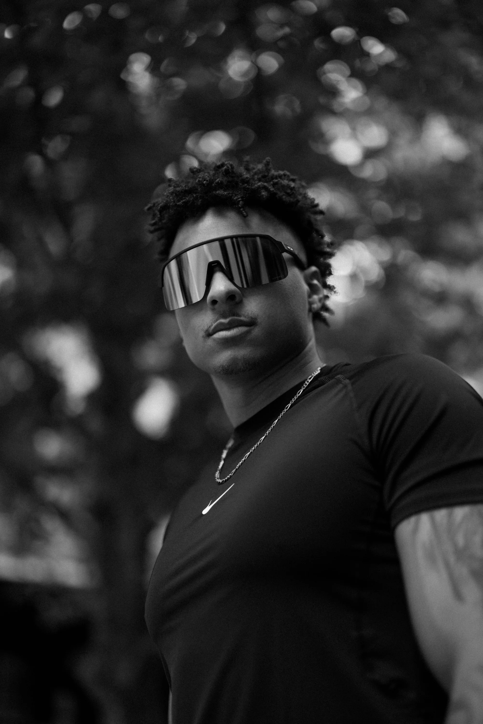 A young person wearing a Nike shirt, chain necklace, and reflective sunglasses, looking to the side with a blurred outdoor background.