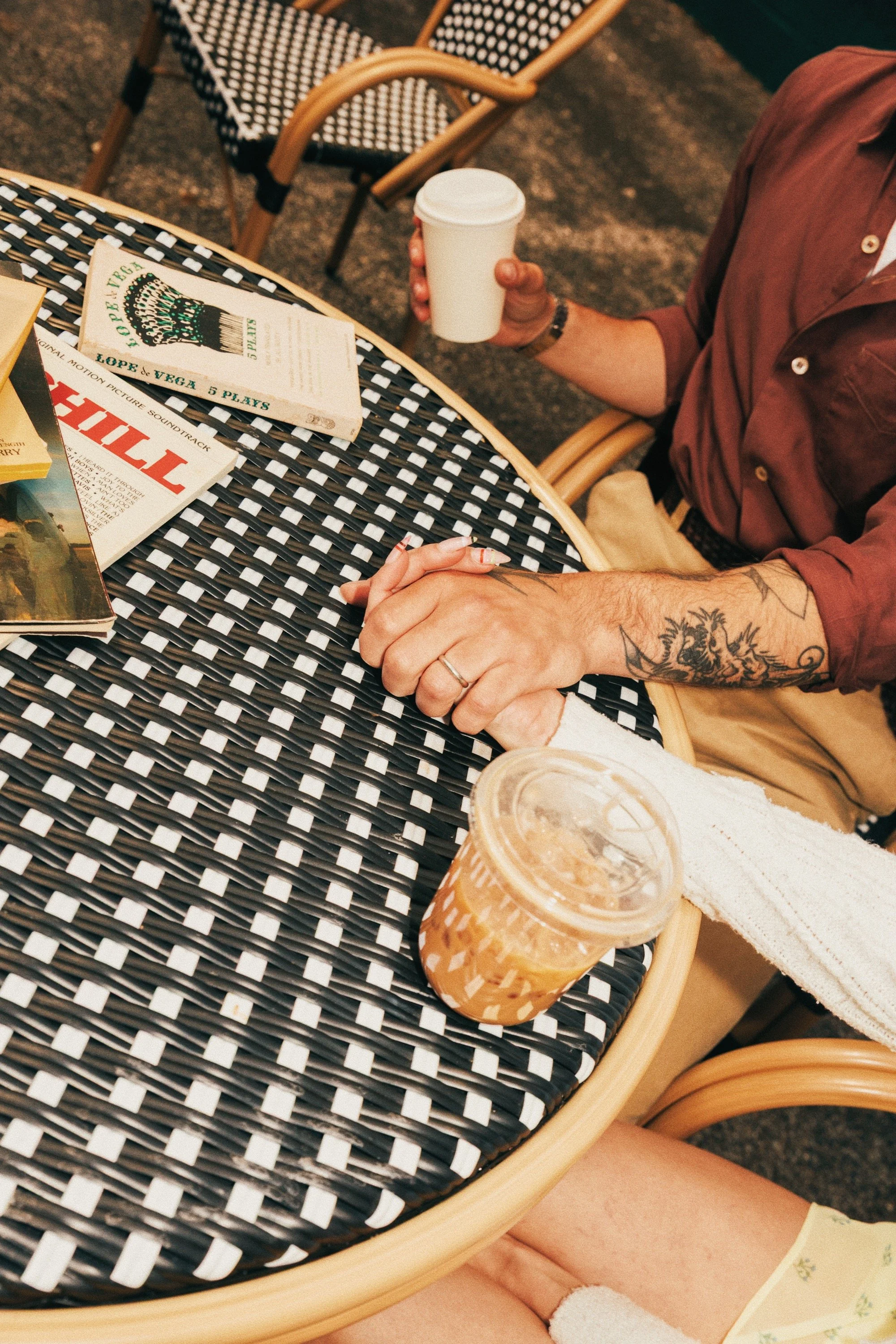 A person sitting at a black and white woven outdoor table, holding a white coffee cup. There are various magazines on the table, a cold beverage in a plastic cup with a straw, and other guests' feet are visible. The setting appears to be outdoors at 