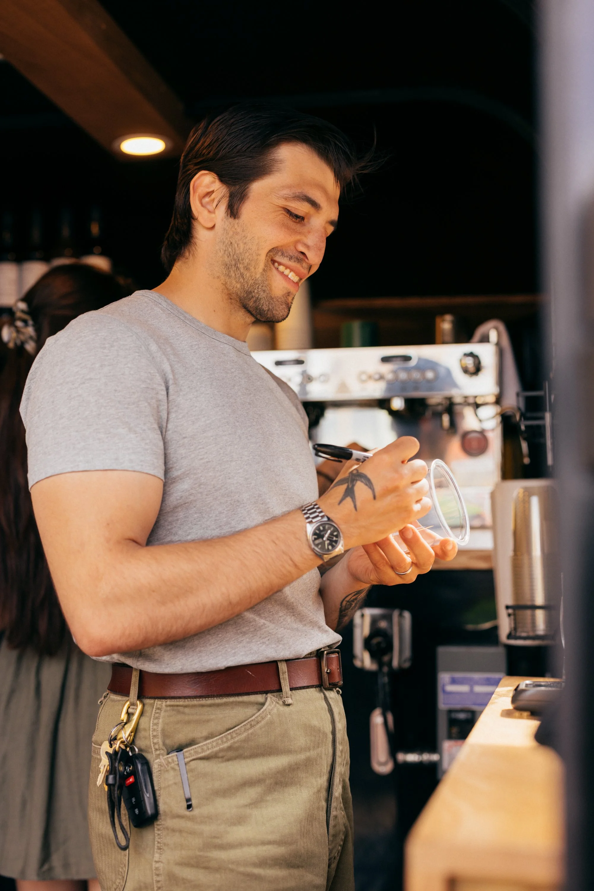A man with dark hair, a beard, and tattoos, wearing a gray t-shirt, is smiling and looking down at his phone while standing at a counter in a coffee shop or cafe. He has keys and a lanyard hanging from his belt. Cafe Stai Manso 