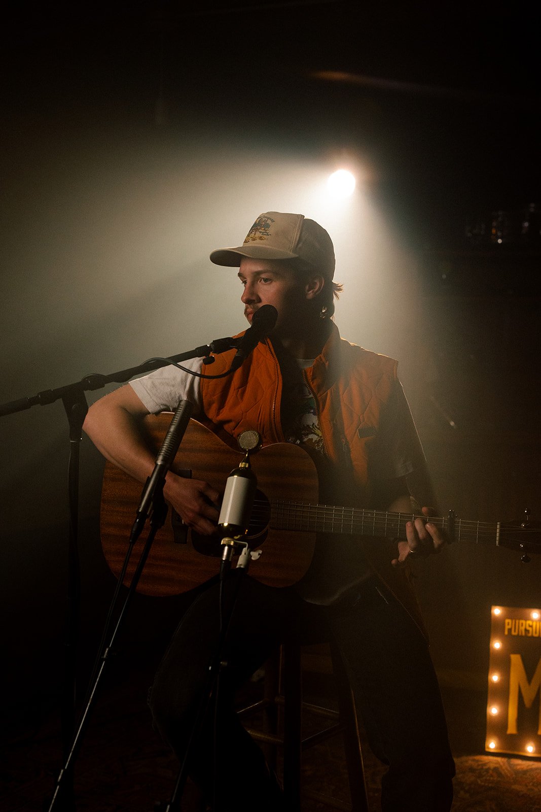 A young man wearing a beige baseball cap and an orange vest is sitting on a stool, playing an acoustic guitar and singing into a microphone in a dimly lit setting with a spotlight behind him.