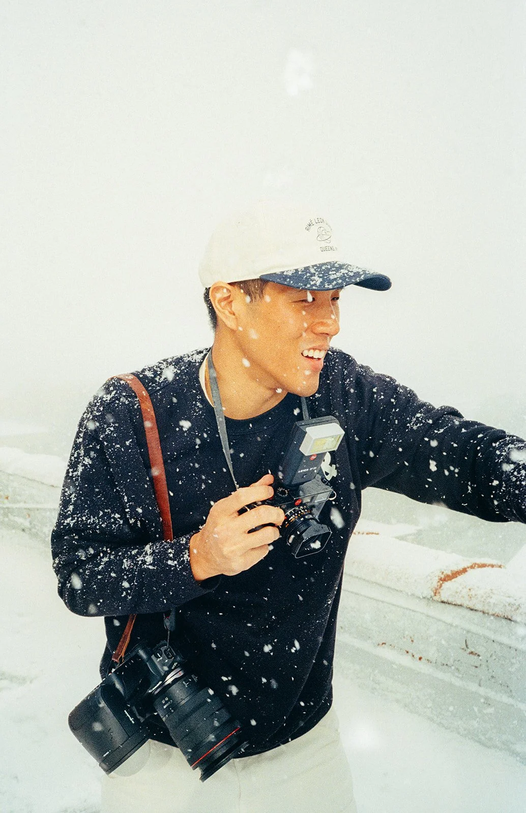 A person wearing a white cap, black sweatshirt, and beige pants is smiling and holding a camera in a snowy environment with snow falling around them.
