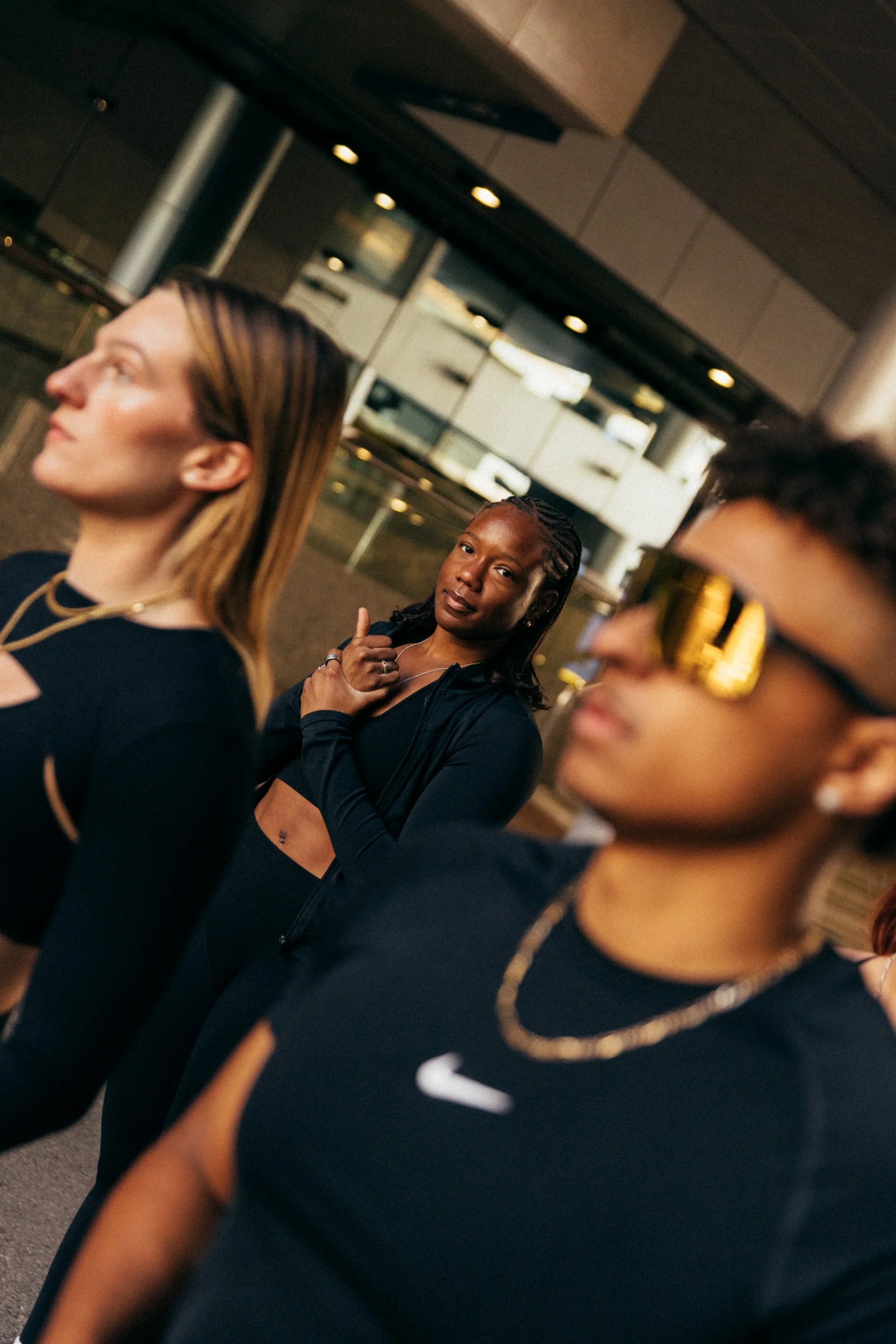 Three women wearing black outfits, standing in a modern indoor setting with glass walls and ceiling lights.