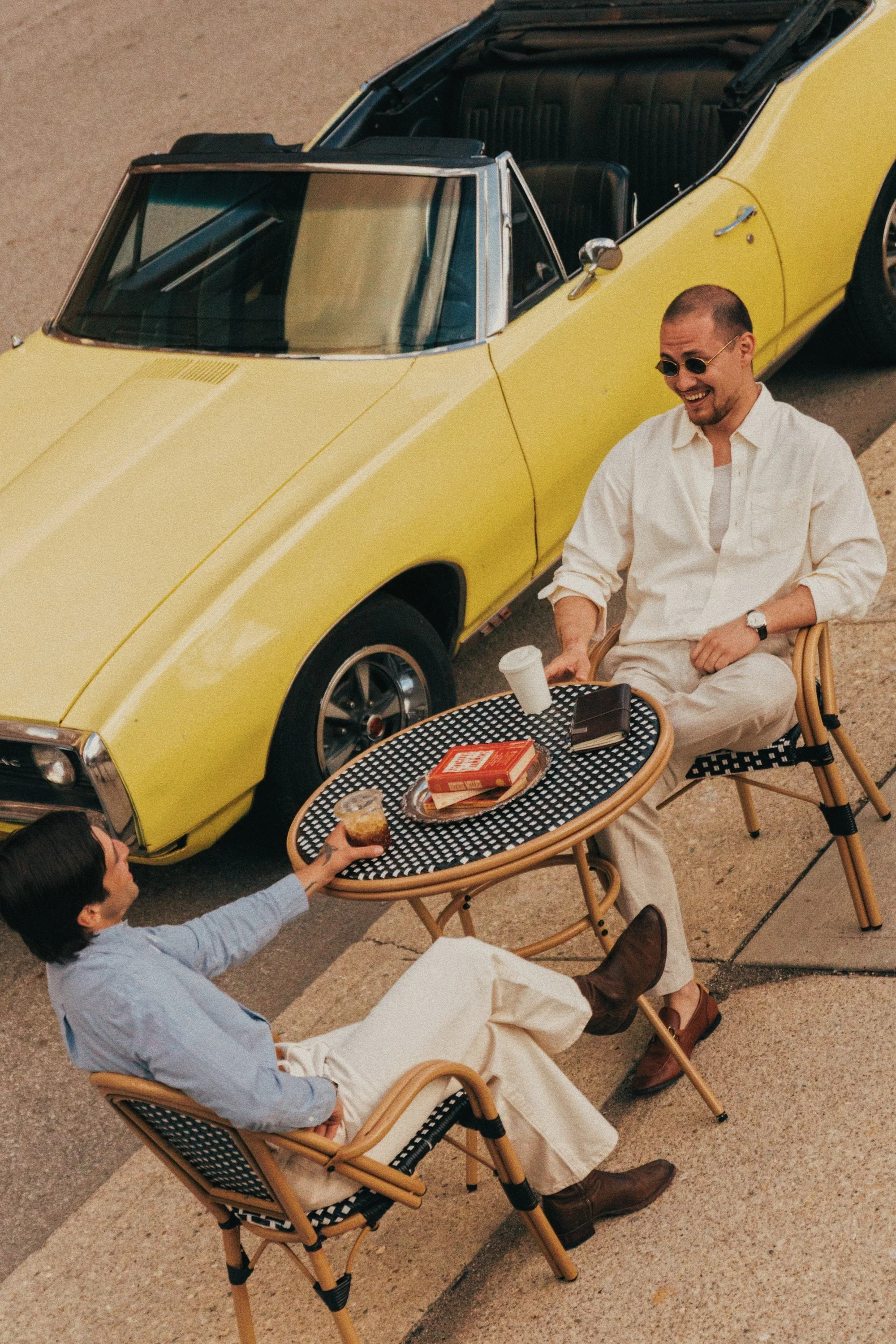 Two men sitting at a small round outdoor table having coffee, with one man passing a pastry to the other. A yellow vintage convertible car is parked nearby.