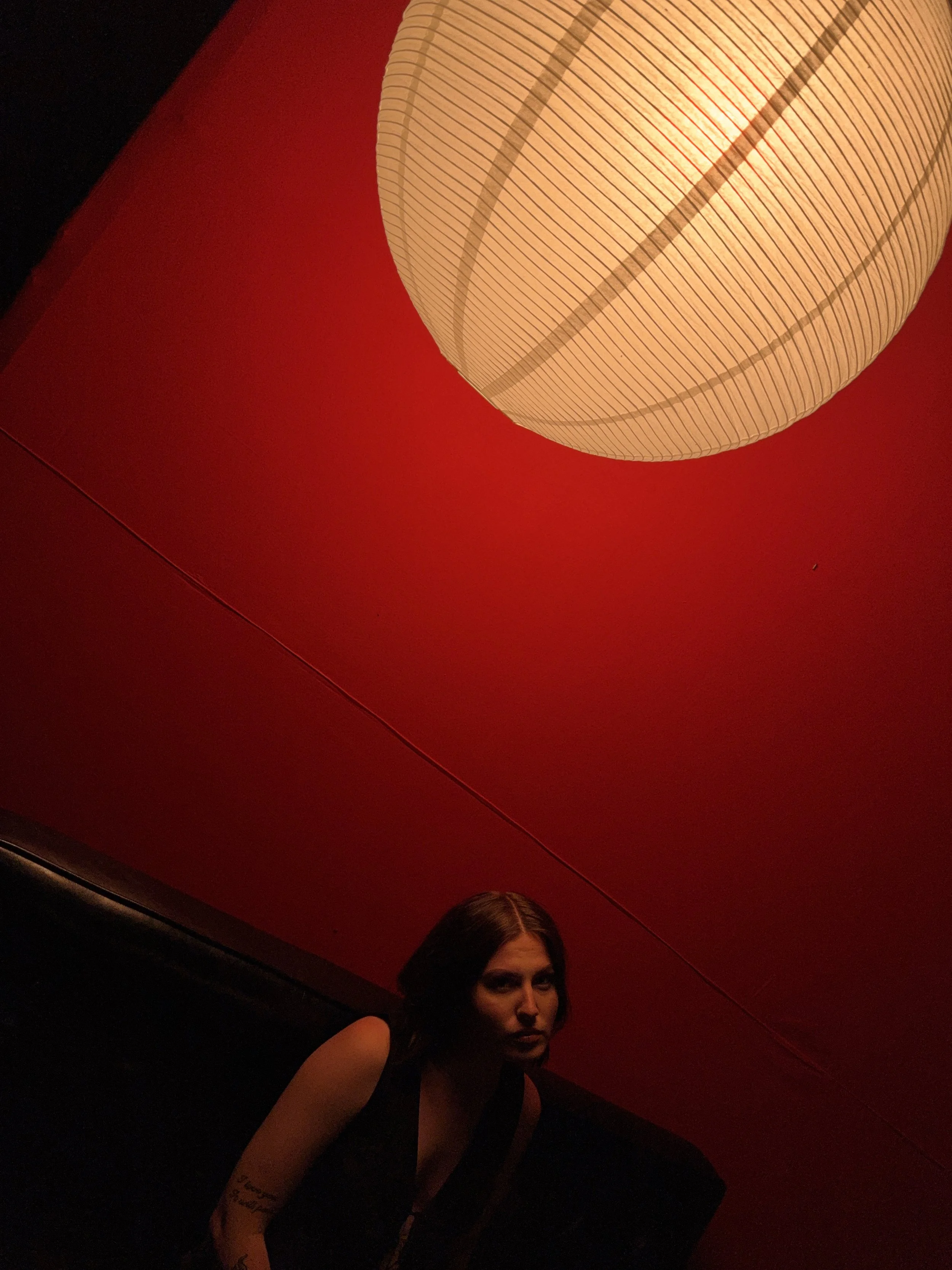 A woman with dark hair and tattoos on her arm sitting against a black and red wall illuminated by a large hanging paper lantern.