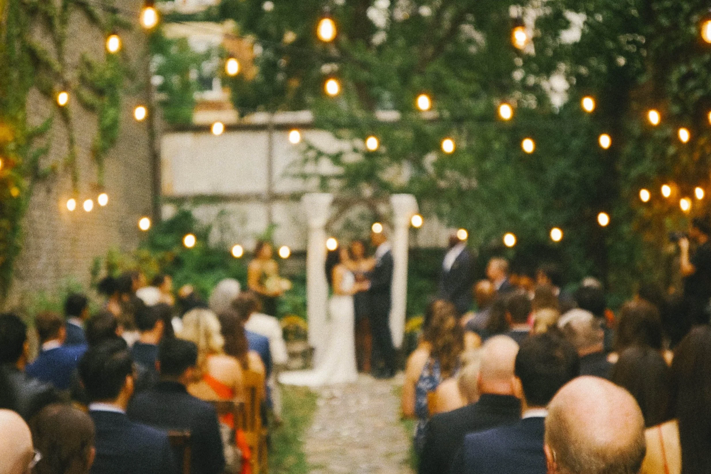A blurred outdoor wedding ceremony with guests seated on either side of a central aisle, a couple standing under a white arch, and string lights hanging overhead.