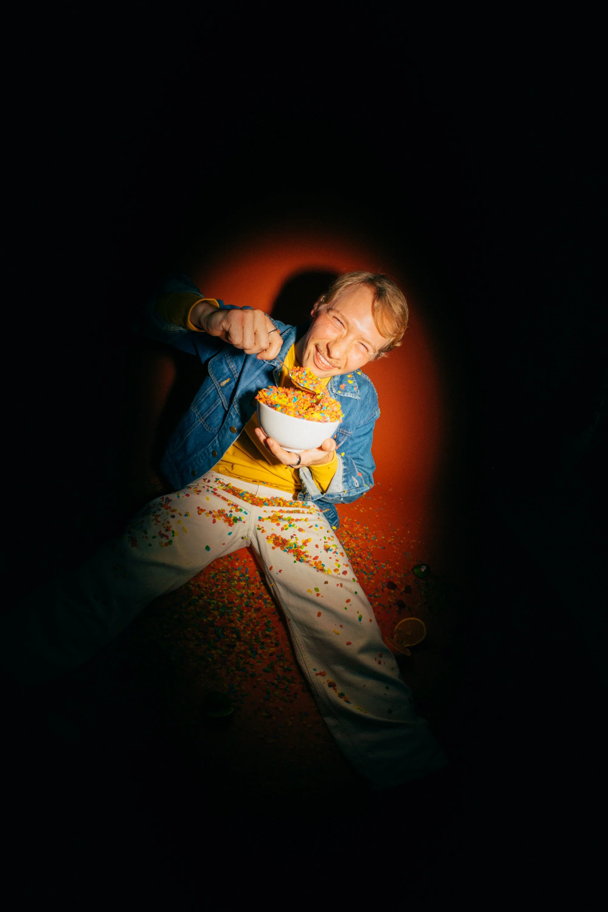 A young man with light brown hair smiling and holding a white bowl of colorful sprinkles, sitting on the floor covered with sprinkles, with a dark background and a warm glow behind him. Cafe Stai Manso