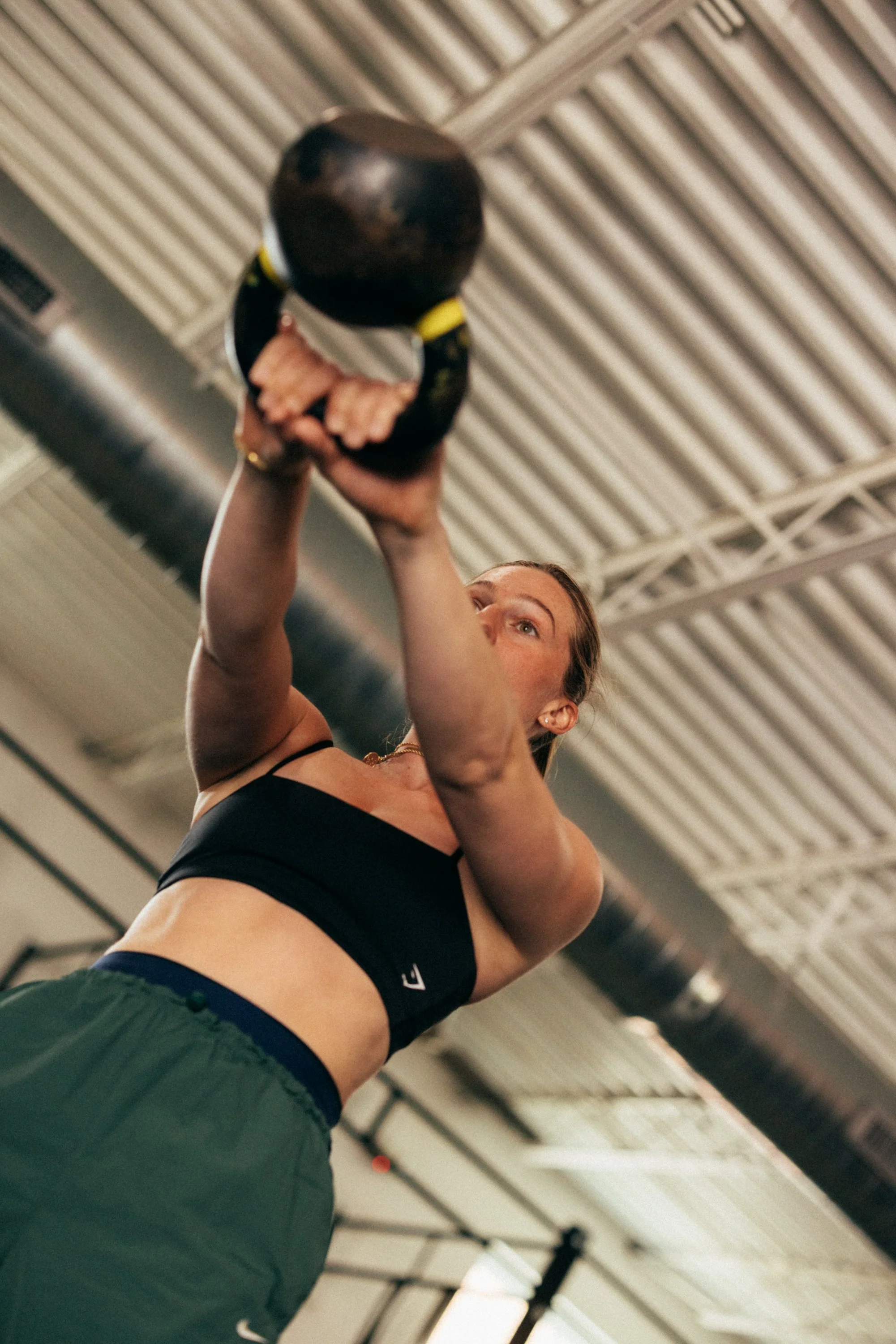 A woman in athletic clothing lifting a kettlebell in a gym with a high ceiling and metal beams.