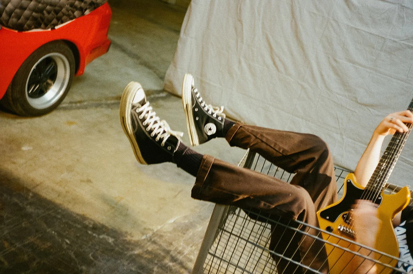 Person lying in a shopping cart holding a yellow electric guitar, wearing black and white sneakers and brown pants.