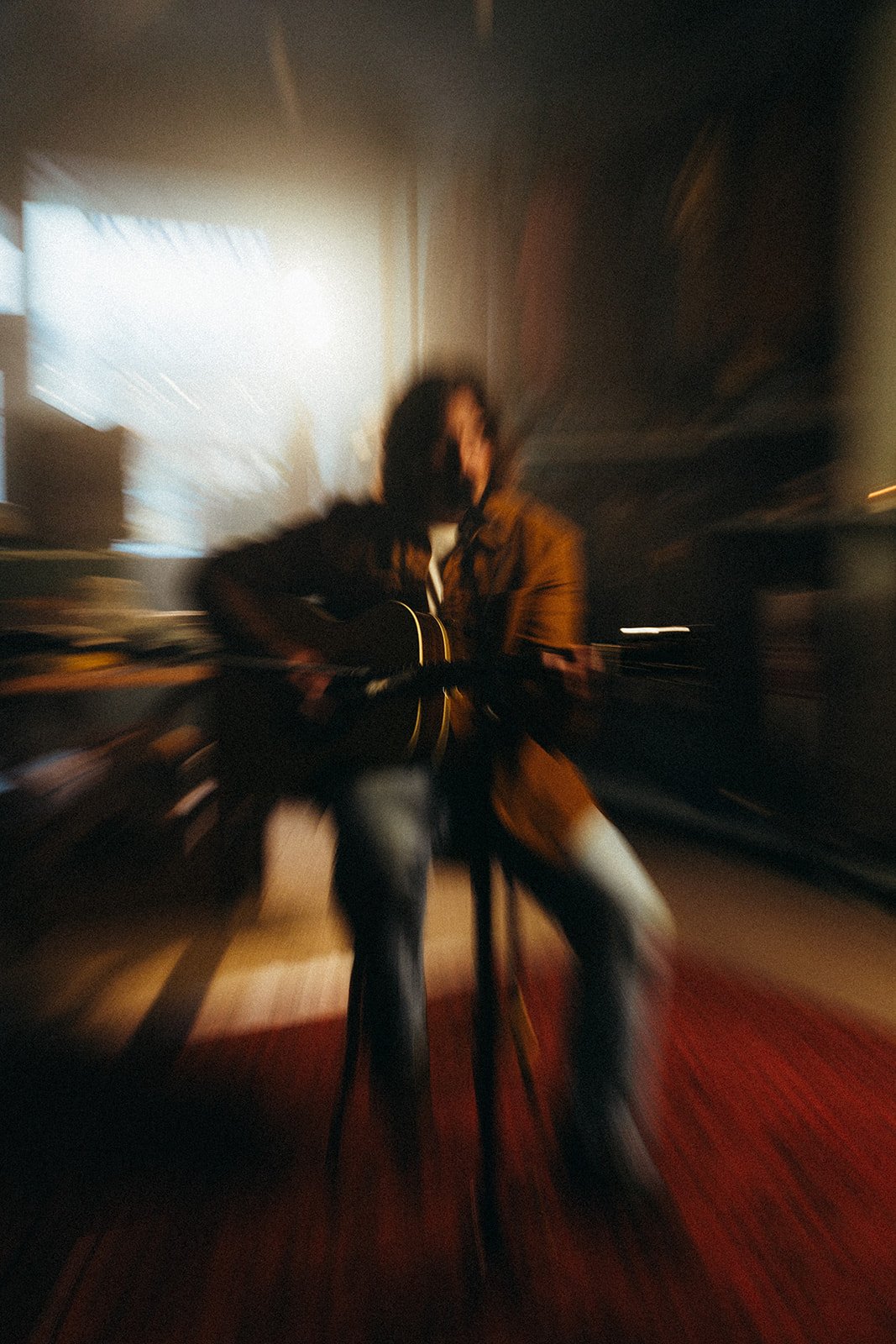 A person sitting on a stool playing an acoustic guitar in a dimly lit room with a window and shelves in the background, with a motion blur effect.