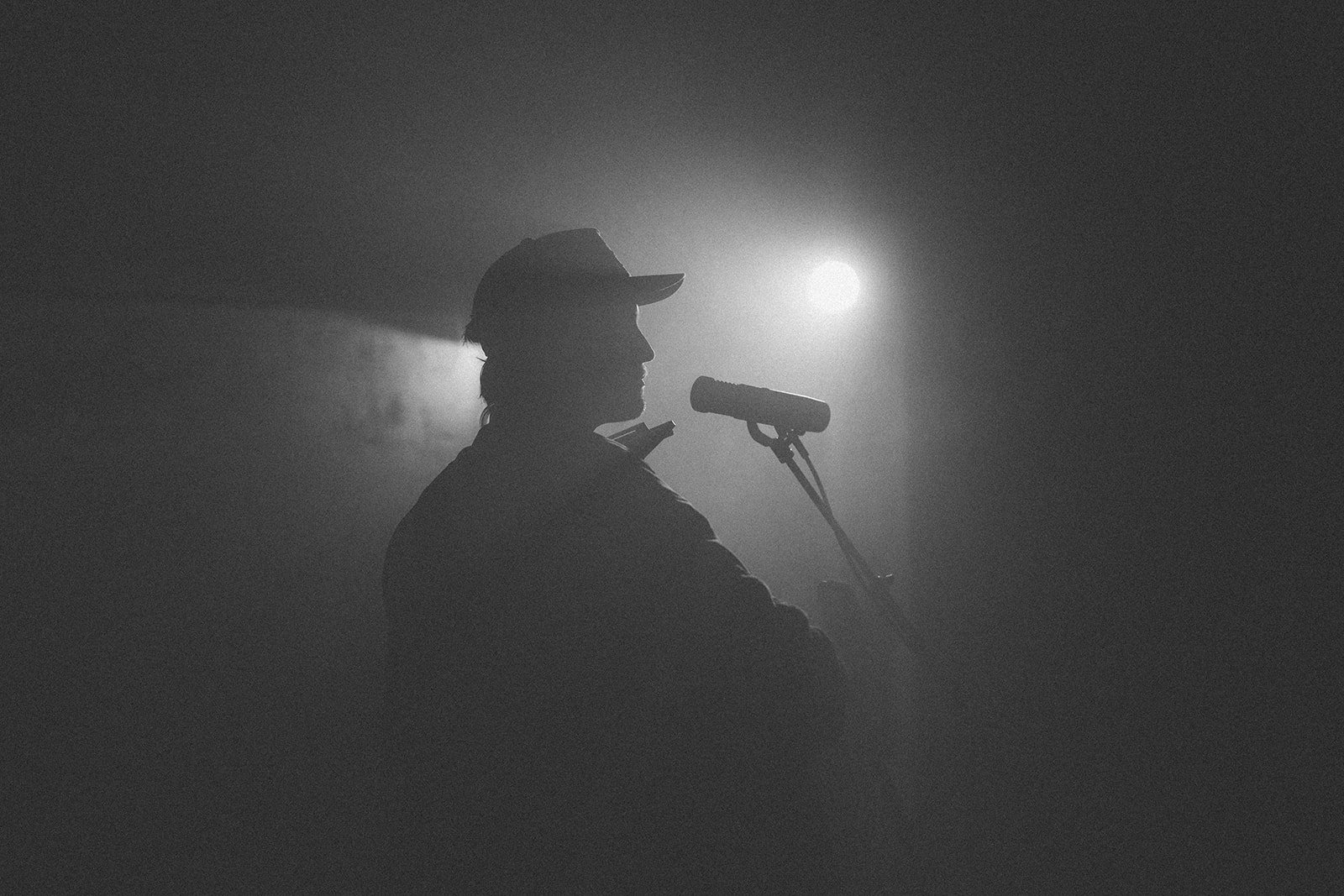 A silhouette of a person wearing a hat, speaking or singing into a microphone, with a bright light behind them creating a dramatic shadow effect, in a dark setting.
