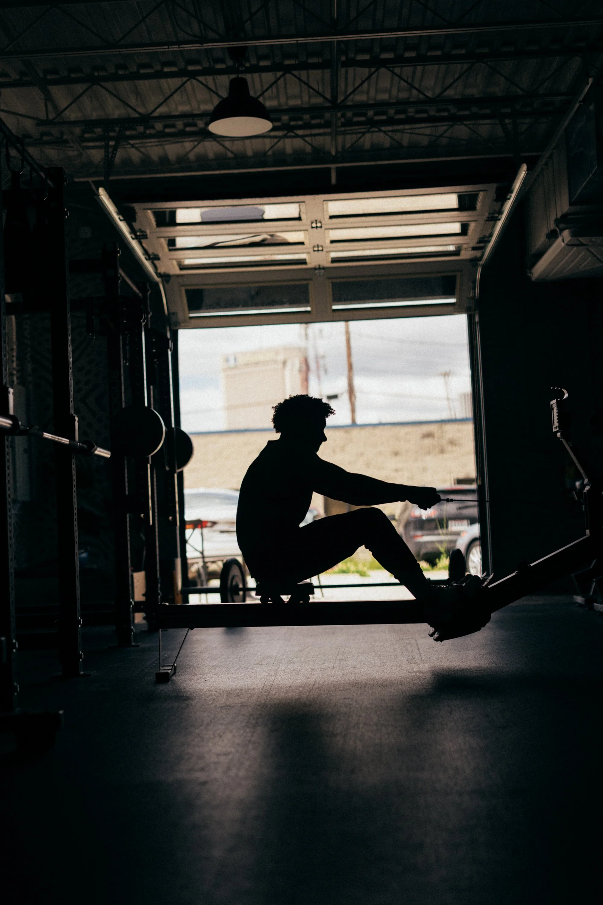 Silhouette of a person stretching or exercising in a gym with an open garage door, outdoor sky and power lines visible in the background.
