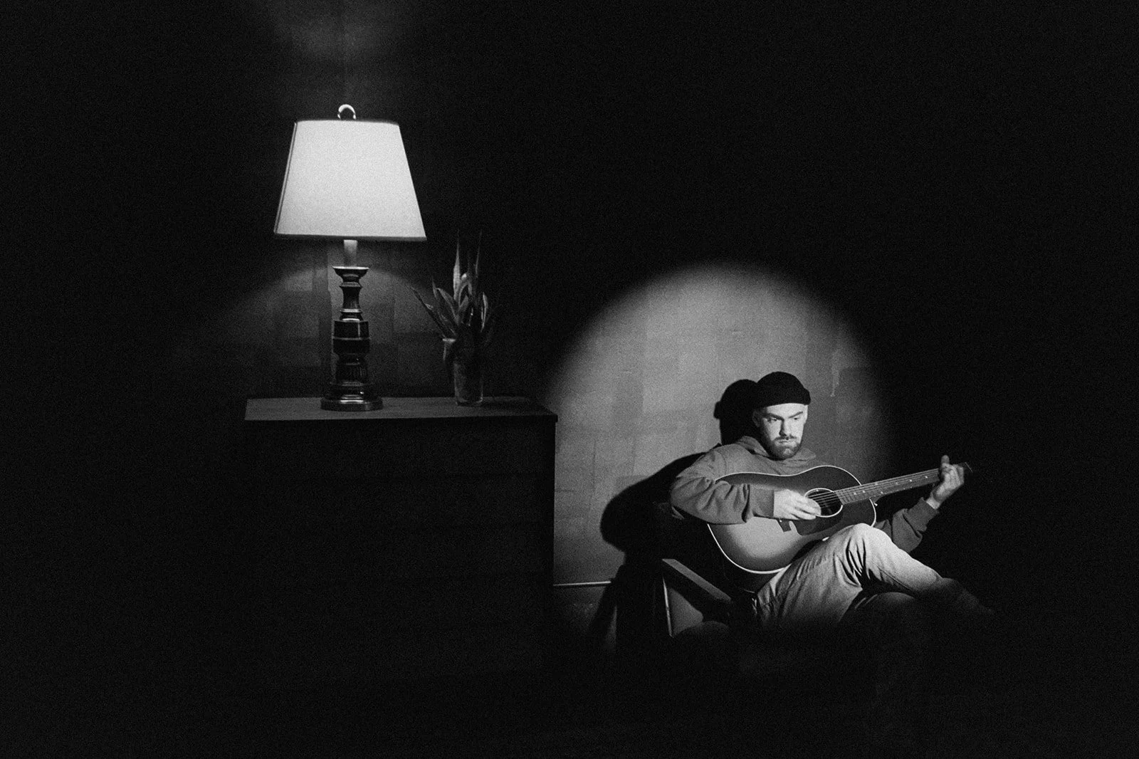 A man sitting on a chair playing an acoustic guitar in a dark room. A lamp on a table beside him illuminates part of the room, casting light on a vase with tall plants.