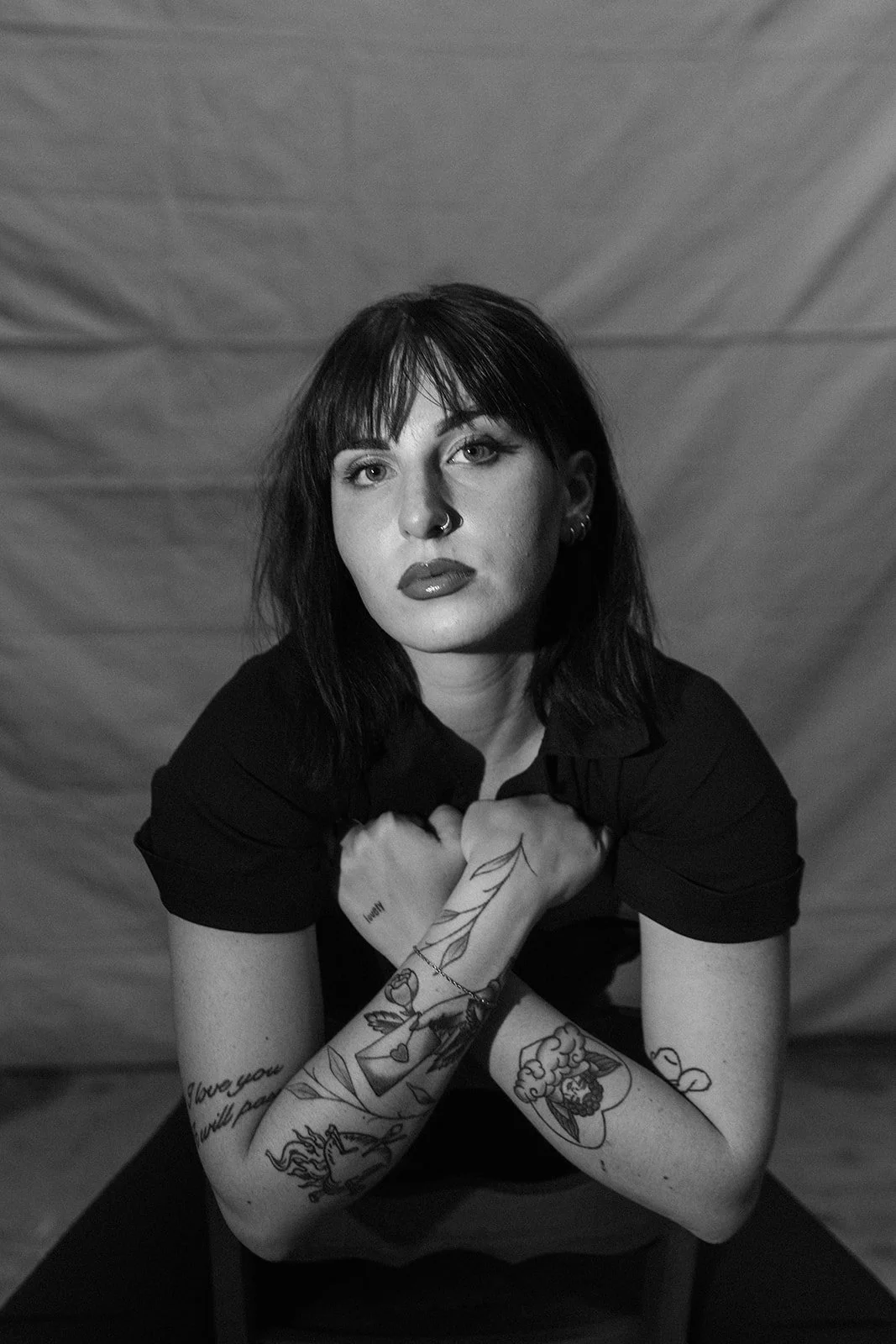 Black and white portrait of a woman with shoulder-length dark hair, tattoos on her arms, and wearing a dark shirt, looking directly at the camera with a serious expression.