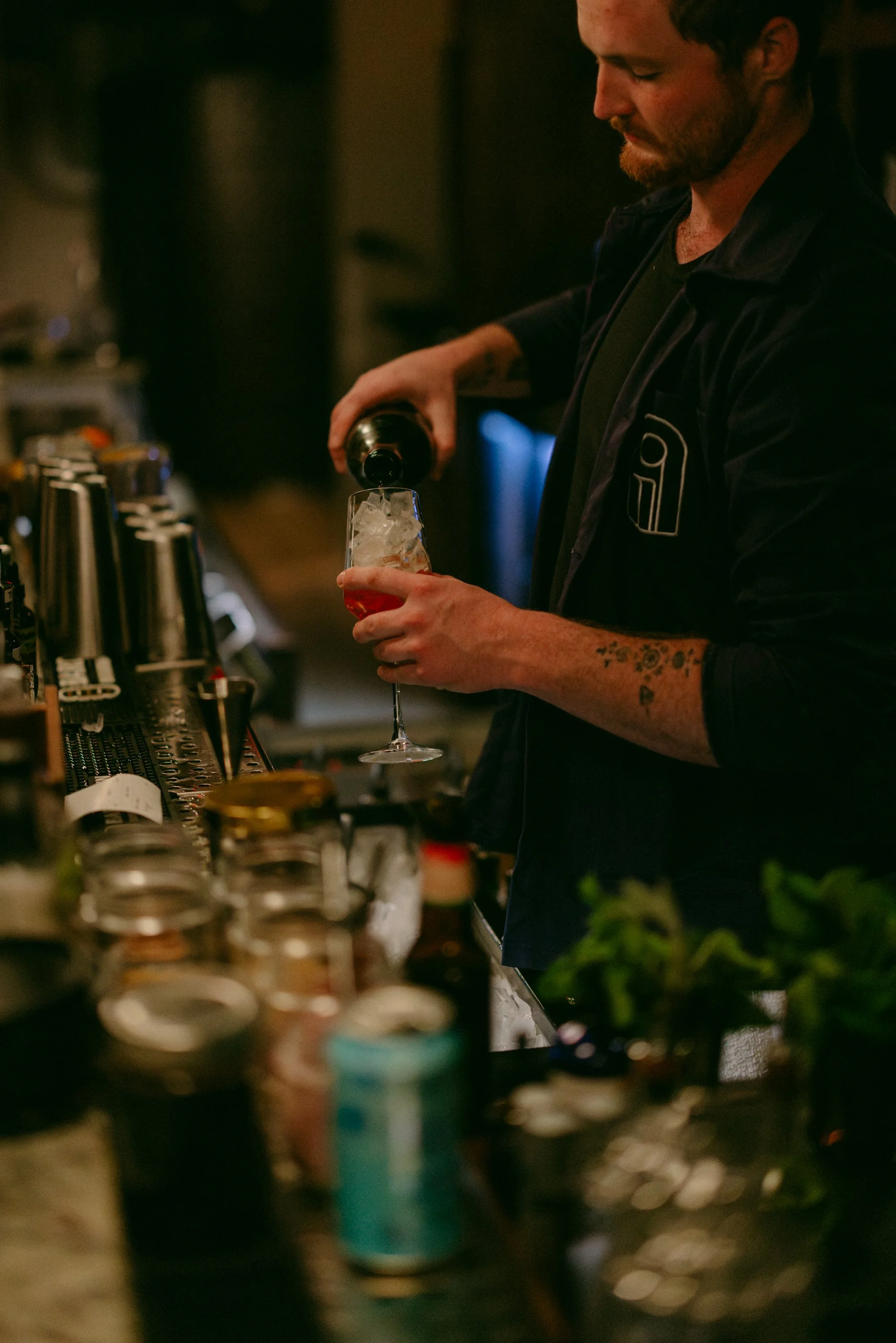 A man with a beard and tattoos on his arm pours a drink from a bottle into a glass filled with ice in a dimly lit bar or restaurant setting.