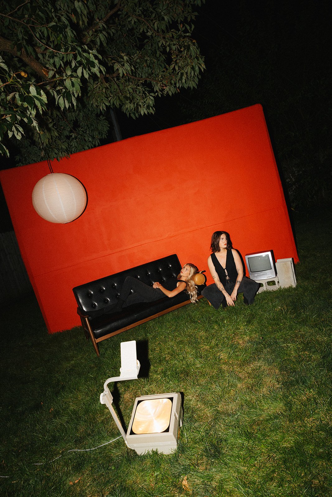 Two women in black clothing outdoors at night, with one sitting on a black couch and the other squatting on the grass, in front of a large red backdrop, surrounded by a tree, a white paper lantern, a small vintage television, and a portable spotlight