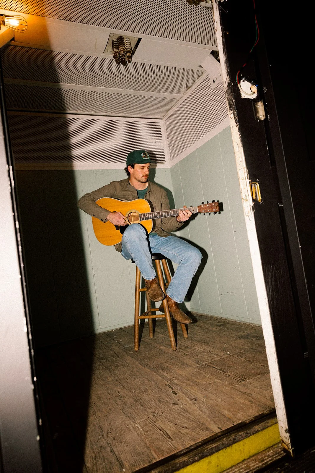 A man wearing a green cap, brown jacket, and jeans is sitting on a wooden stool in a small, plain room playing an acoustic guitar.