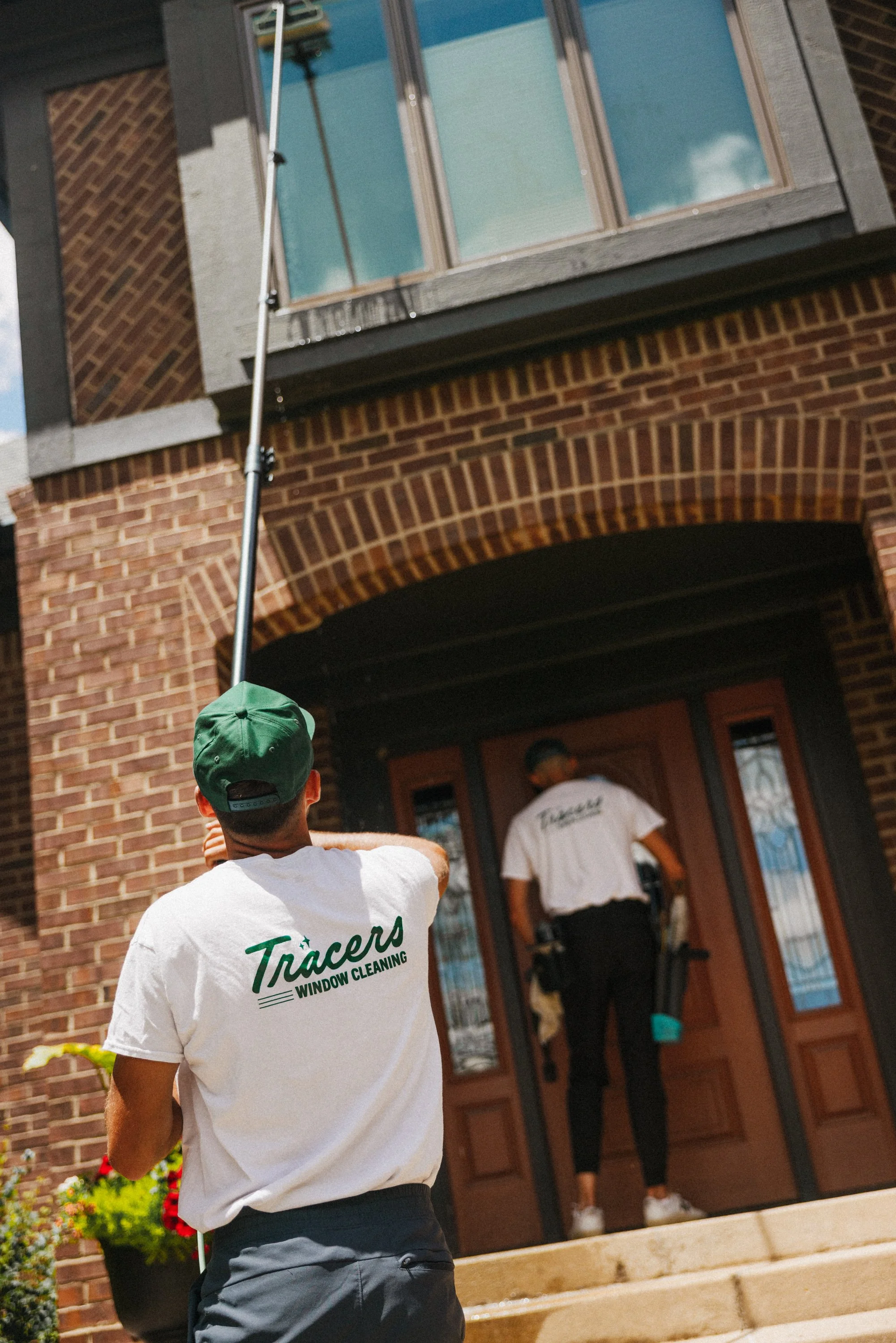 A man with a green cap and white Tracers window cleaning t-shirt is holding a long pole, reaching towards a second-floor window of a brick building as a woman in a white T-shirt and black pants stands at the door in the background.
