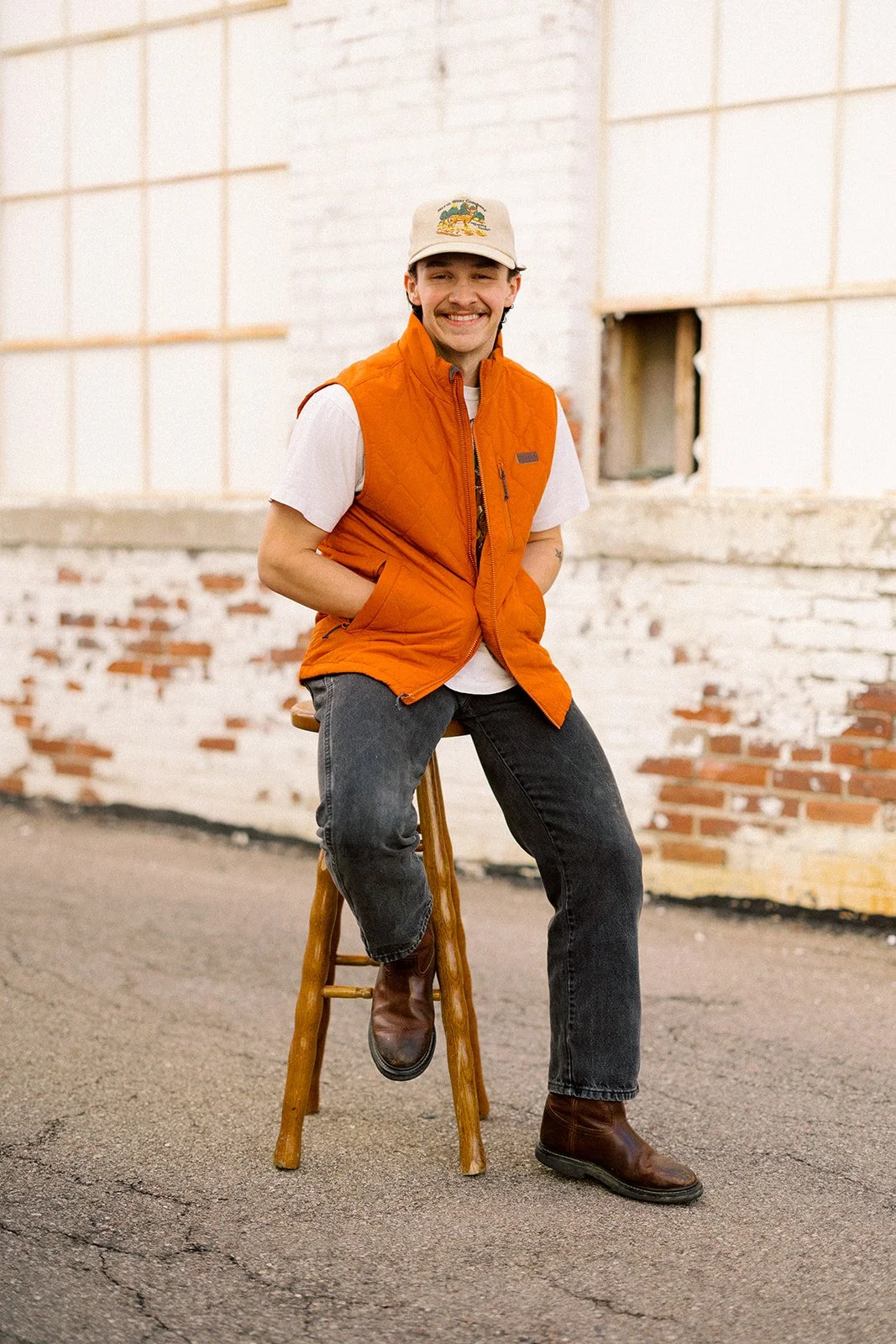 A young man sitting on a wooden stool outdoors, wearing a beige cap, orange vest, white t-shirt, black jeans, and brown boots, smiling at the camera against a white brick wall.