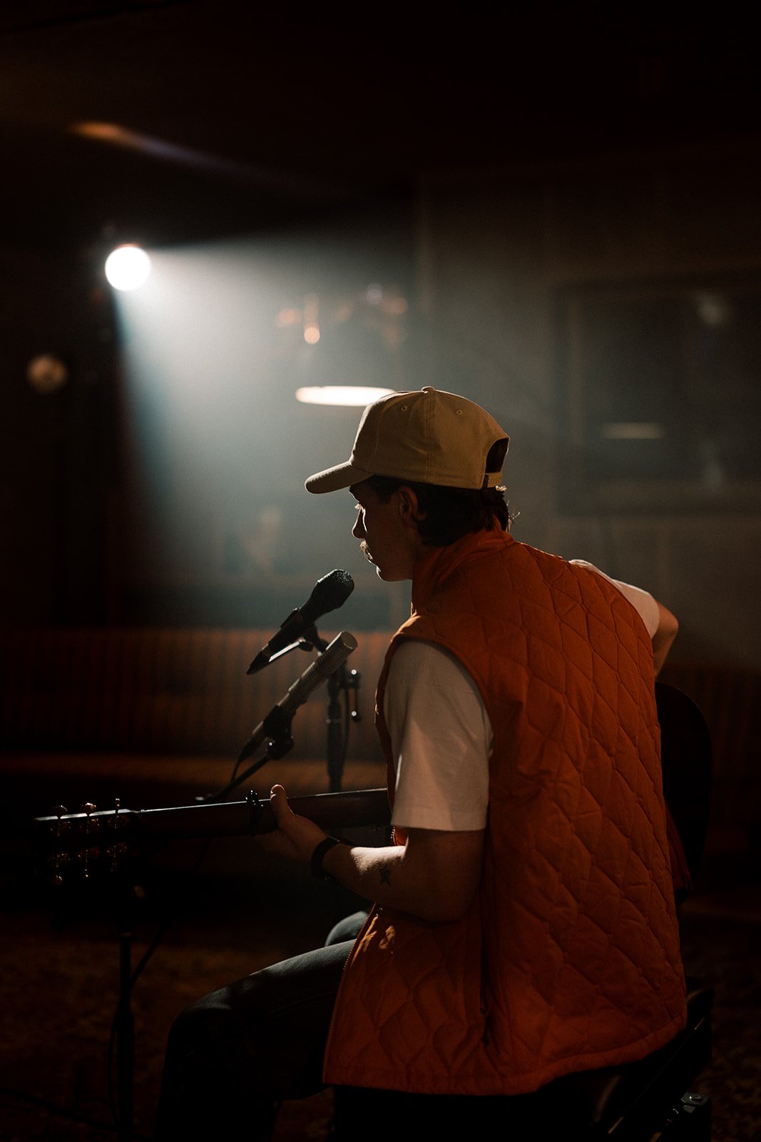 A person wearing a beige cap and orange quilted vest playing an acoustic guitar and singing into a microphone in a dimly lit room.
