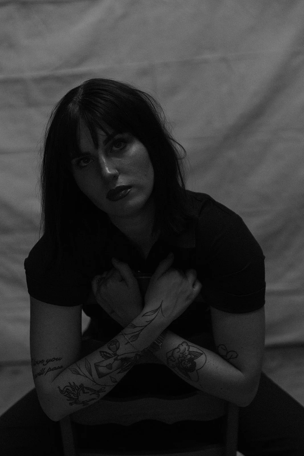 Black and white photo of a woman with dark hair, tattoos on her arms, wearing dark lipstick, looking at the camera with hands crossed over her chest, seated in front of a plain background.