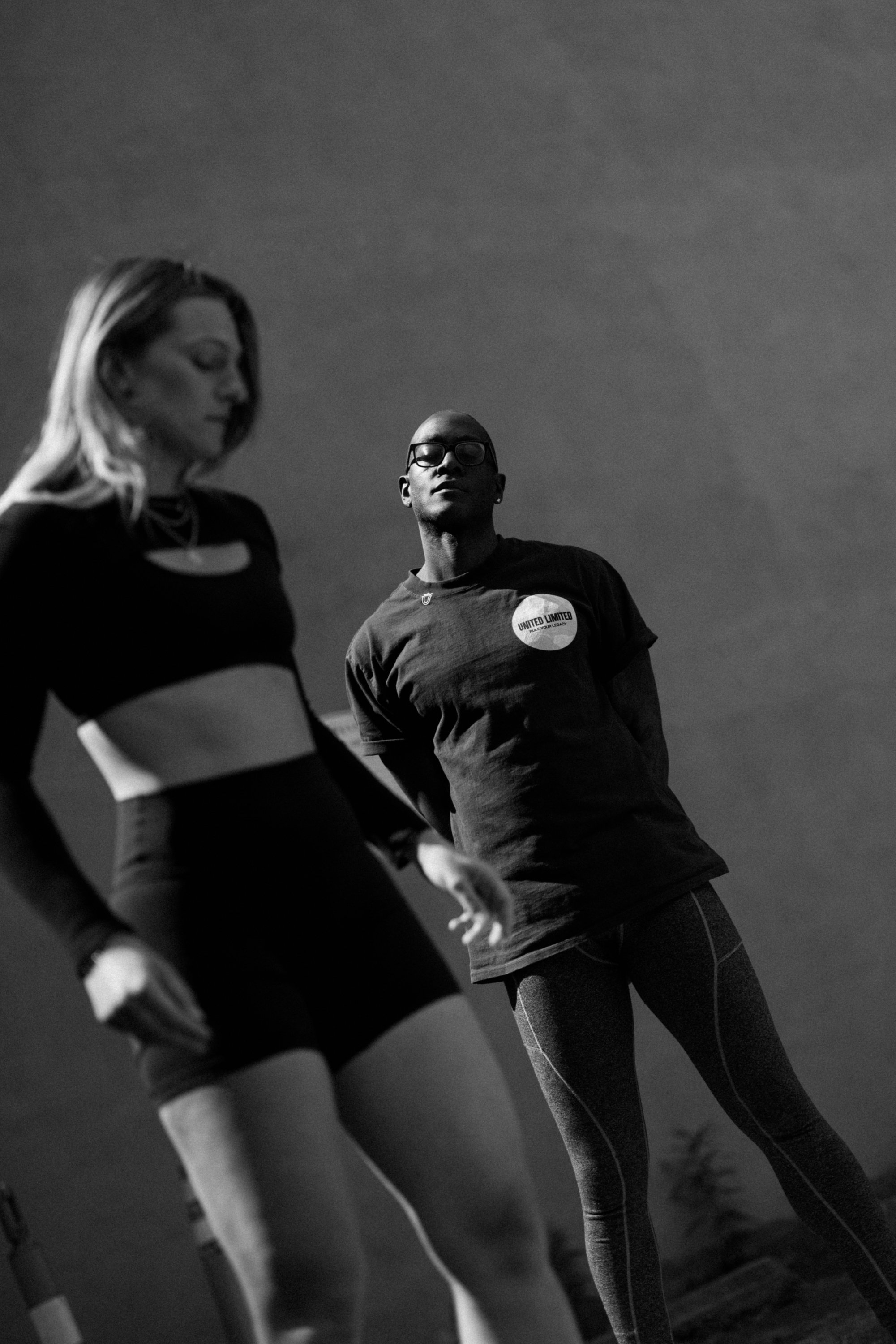 Black and white photo of three women in athletic wear, standing outdoors, under a cloudy sky. Their faces are partially blurred, with one woman in the background looking down, and the others facing outward.