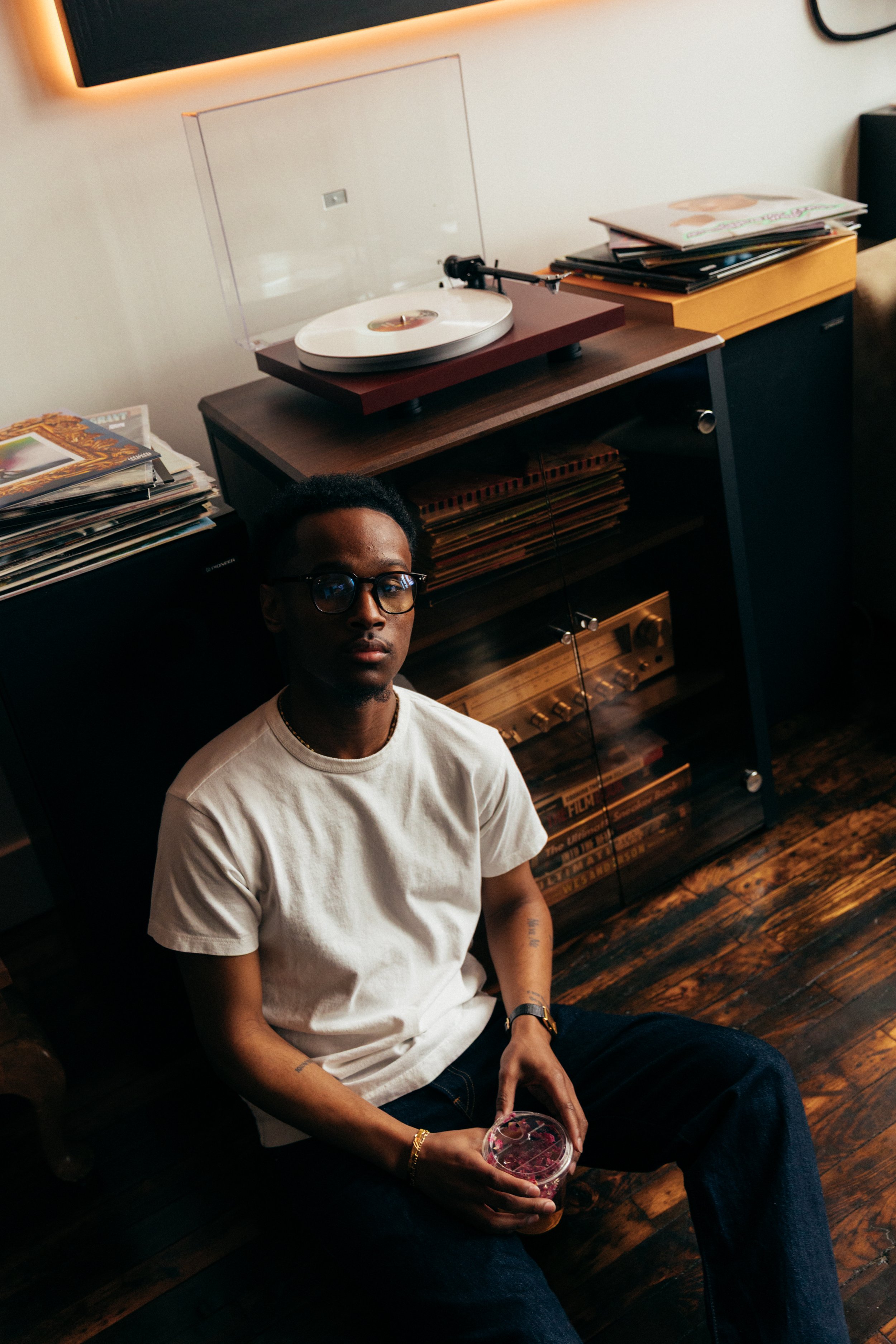 A young man with dark hair and glasses sitting on a wooden floor, holding a plastic cup, in a room with vinyl records, a turntable, and a sound system. Cafe Stai Manso