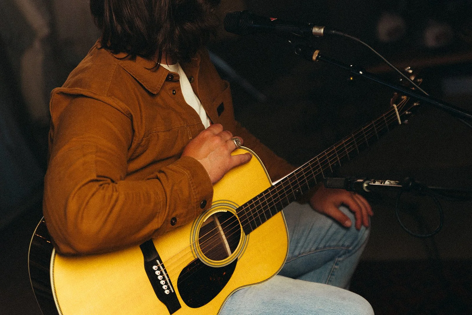 Person playing yellow acoustic guitar while sitting on a dark couch, with microphone in front, wearing a brown jacket and jeans.