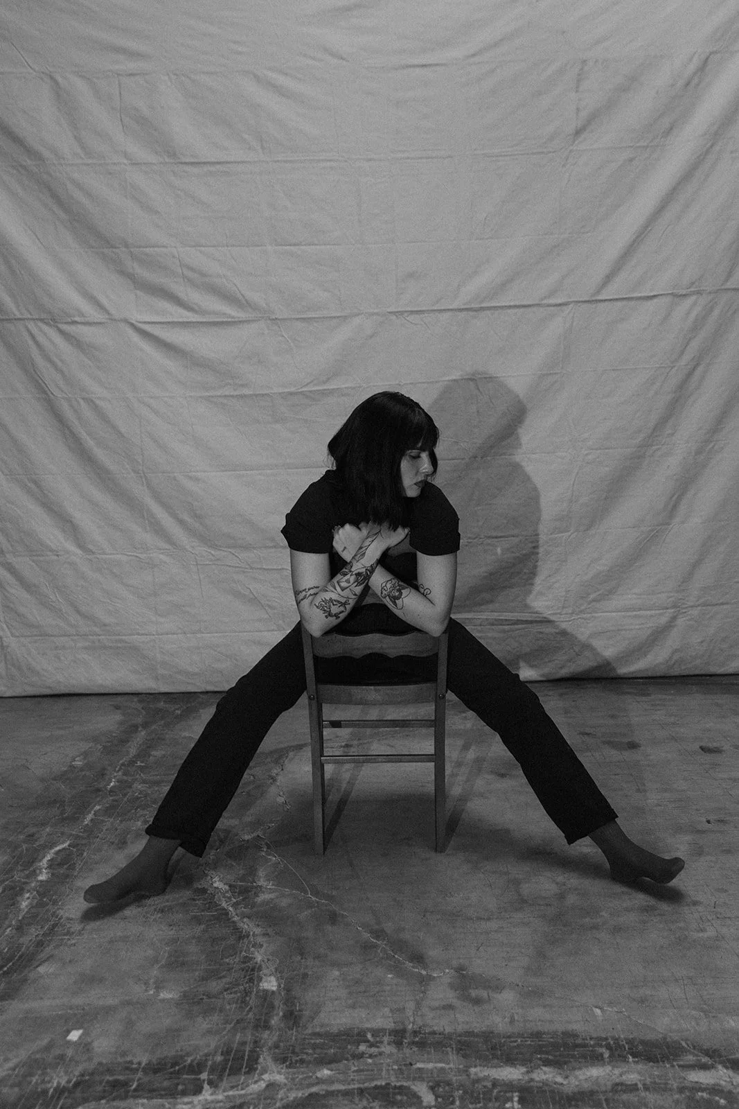 A woman with tattoos, dark hair, wearing a black shirt and pants, sits on a chair with her legs spread wide and her arms crossed over her chest, looking down, in front of a plain backdrop.