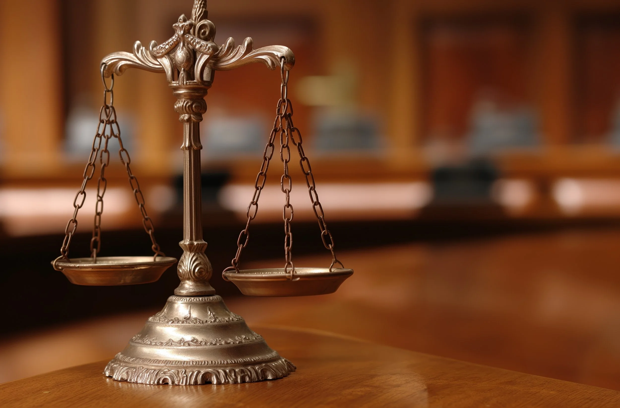 A bronze balance scale on a wooden table, with a traditional courtroom background in the blurred background.