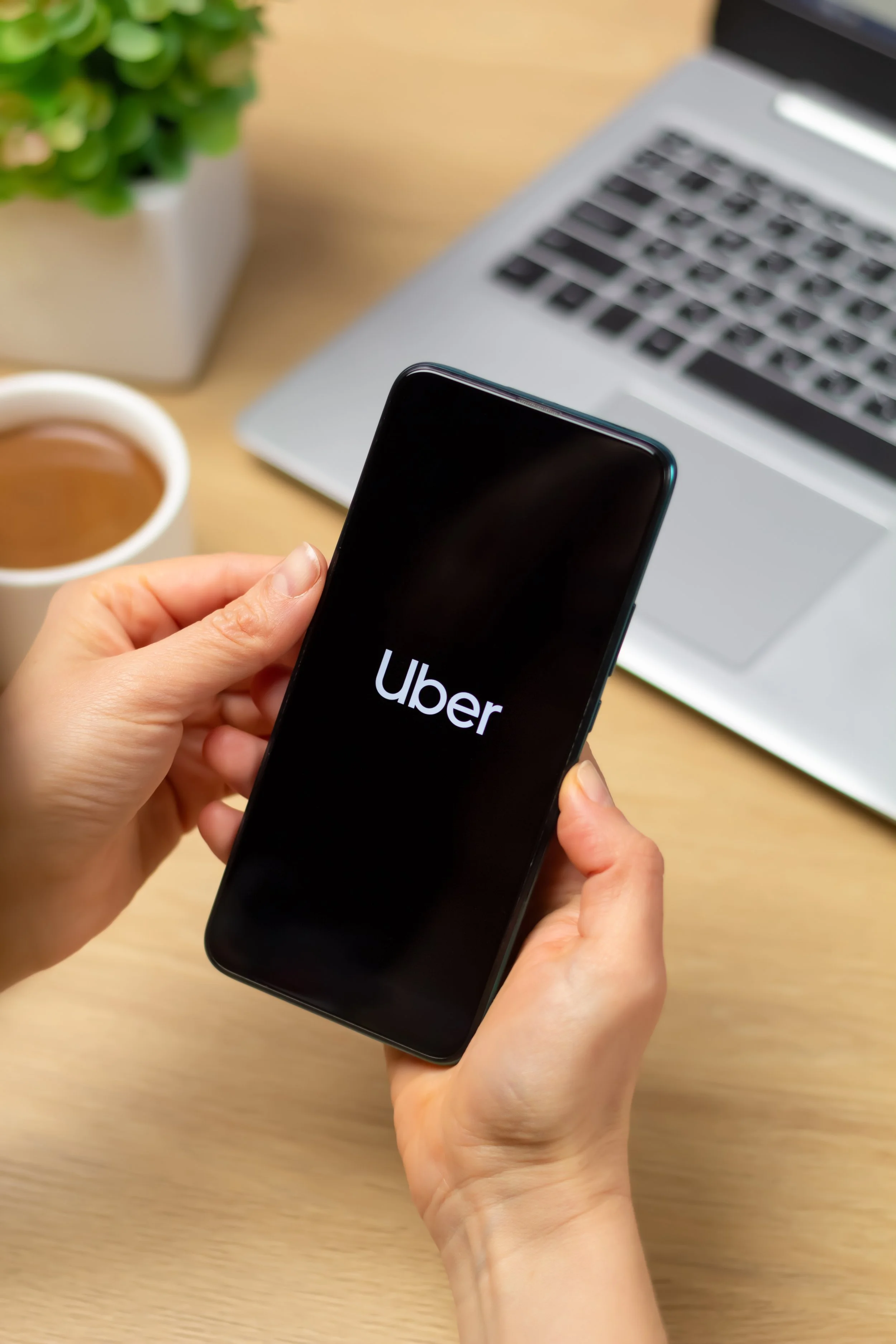 Person holding a smartphone displaying the Uber app logo, on a desk with a laptop, a cup of coffee, and a potted plant.