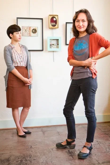 Two women standing in an art gallery with framed artworks on the wall behind them. One woman is looking at the other with her hands clasped in front, while the other woman is standing confidently with one hand on her hip.