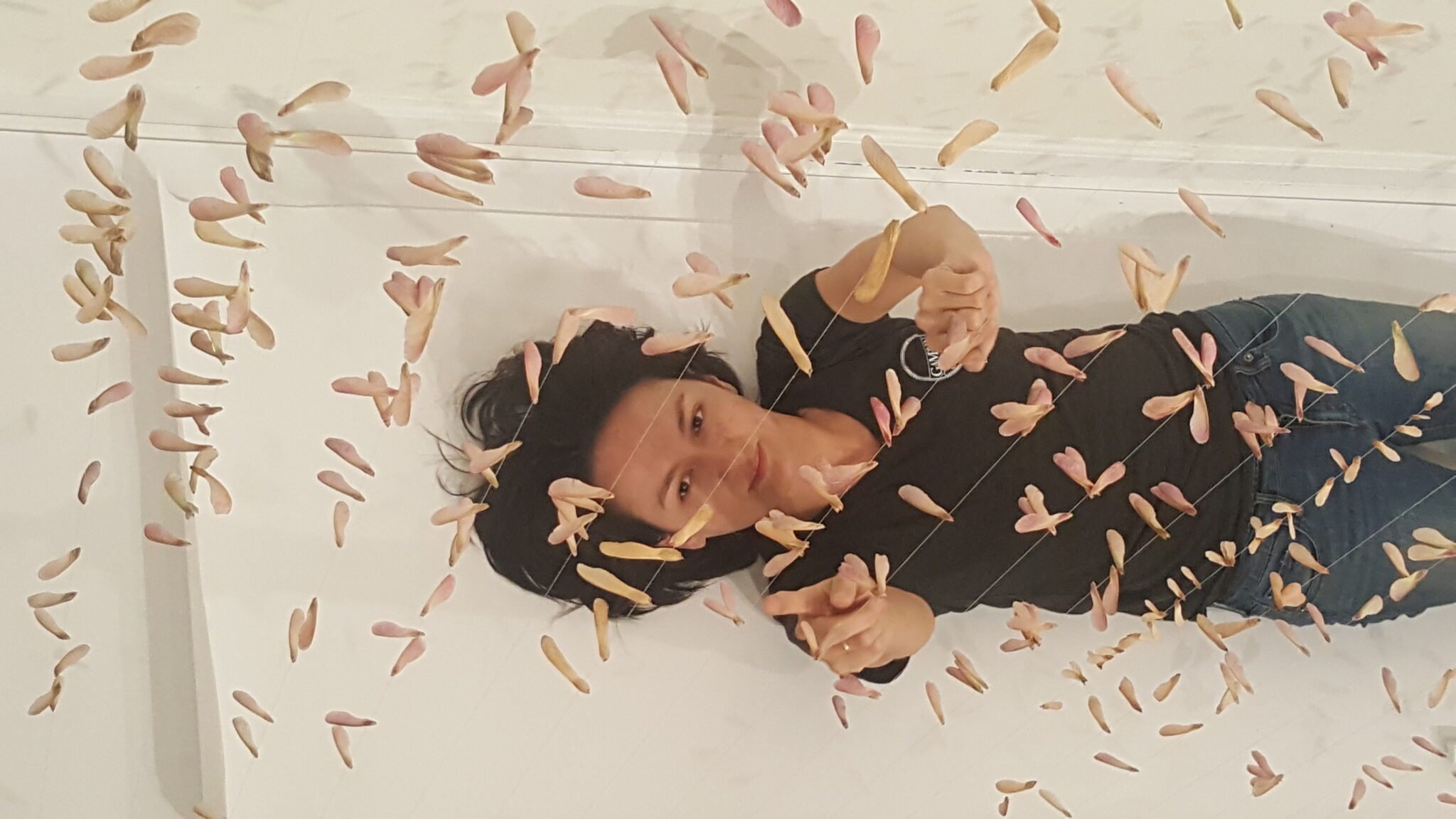 A woman with black hair lying on the floor surrounded by hanging pink and beige flower-like decorations, looking at the camera and pointing.