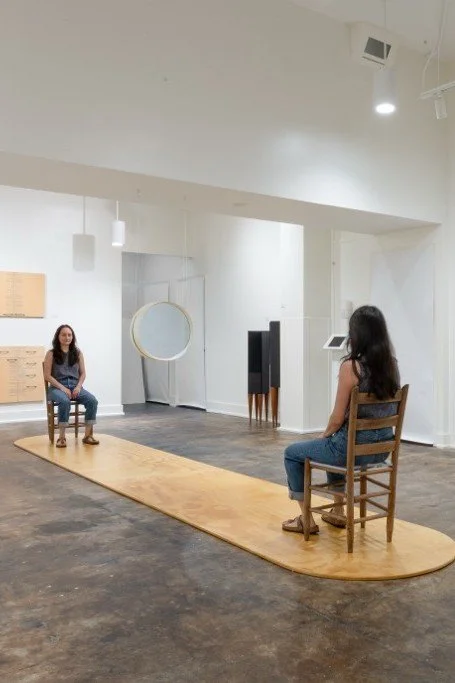 Two women in a minimalist studio, one sitting on a chair on a wooden platform, and the other seated on a chair facing her. The space has white walls, modern lighting, and a mirror with a circular frame.