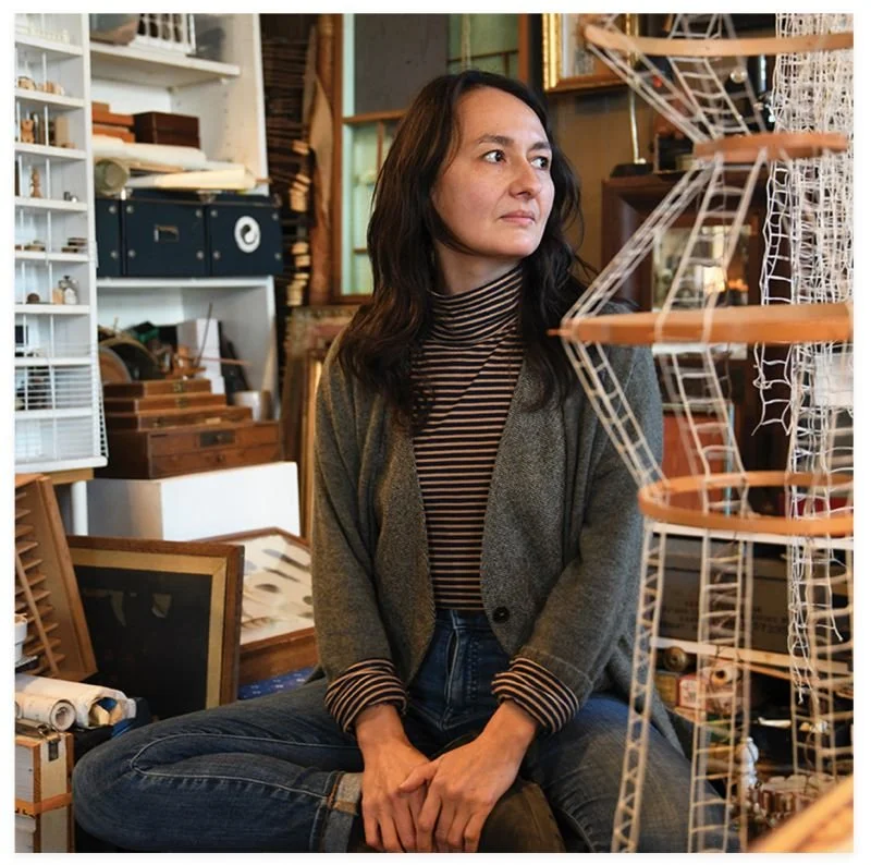 A woman with dark hair wearing a striped turtleneck and gray blazer sitting cross-legged in an antique or craft store, surrounded by various art and craft supplies and models.